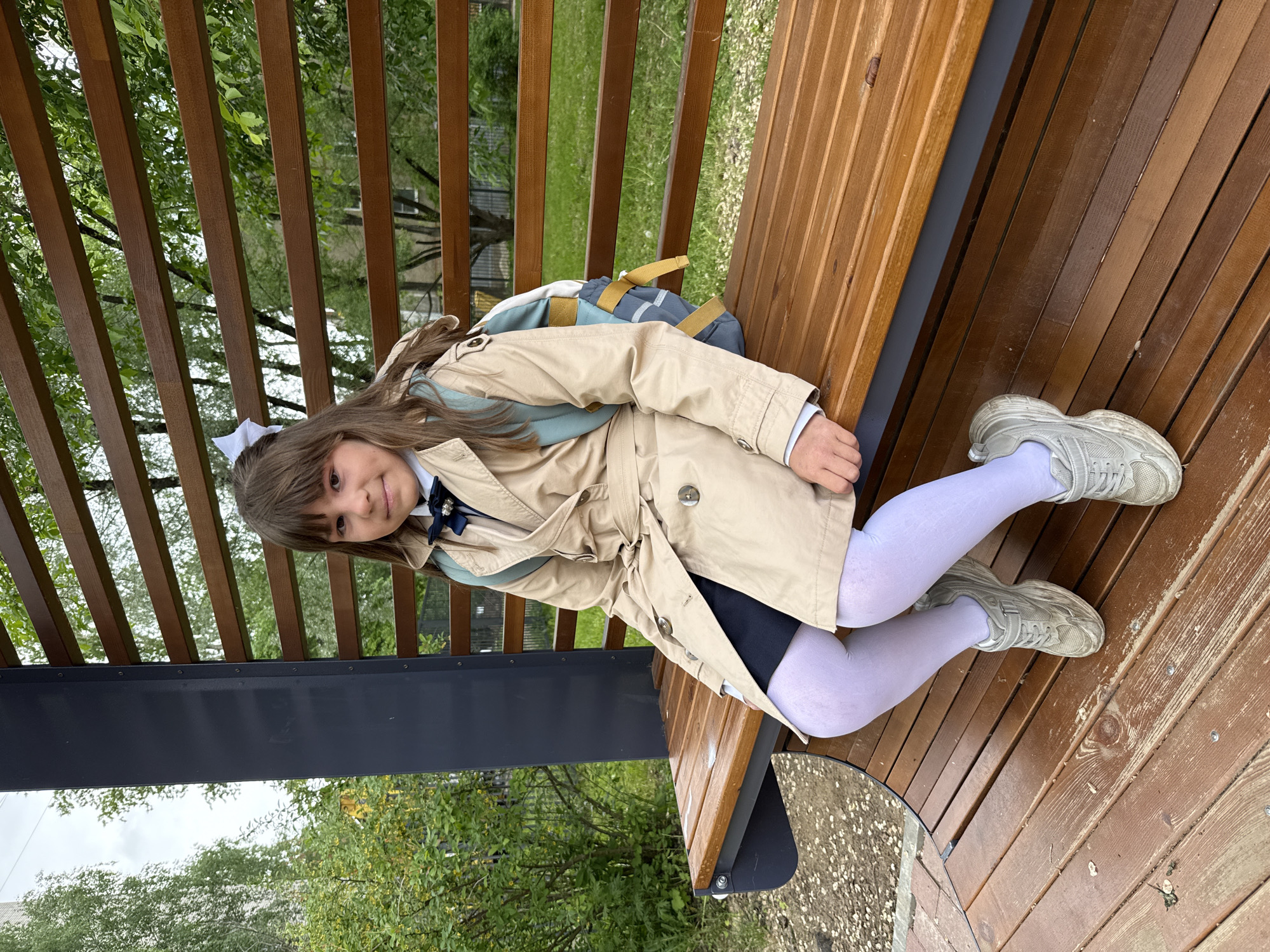 A young girl is sitting on a wooden bench, possibly in a park-like setting. She is wearing a beige trench coat over what appears to be a school uniform with a bow tie and a white hair bow. She also has white tights and gray sneakers. A backpack rests behind her. The bench is constructed with parallel wooden planks, and the area behind it appears to be grassy with trees. The background suggests an outdoor setting, likely in an urban environment. The location, Rusanovo, Russia is specified. The scene is a candid shot of the girl relaxing outdoors.
