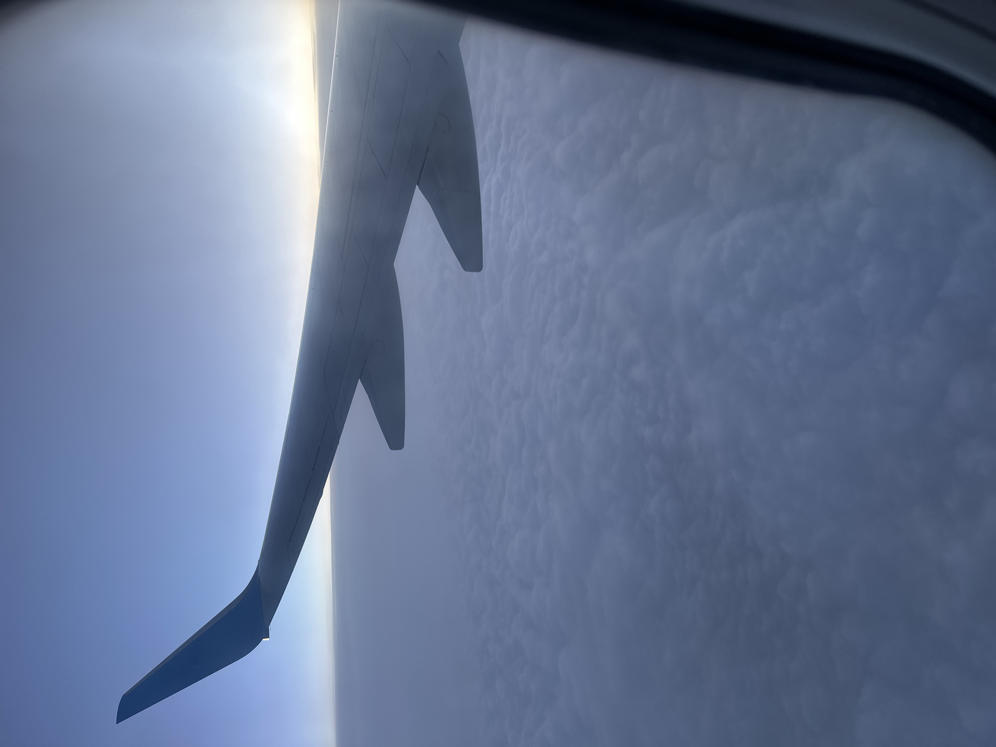 The image shows a view from an airplane window. Part of the airplane wing is visible, with blue and grey coloration. Beyond the wing, there's a dense cloud formation. The presence of the wing suggests the image was taken during a flight. The location (Kitsjanovo, Russia) cannot be confirmed from the image's visual information. There is no visible indication of any people, objects, actions, or interactions. The environment consists of the sky, clouds, and the airplane wing.