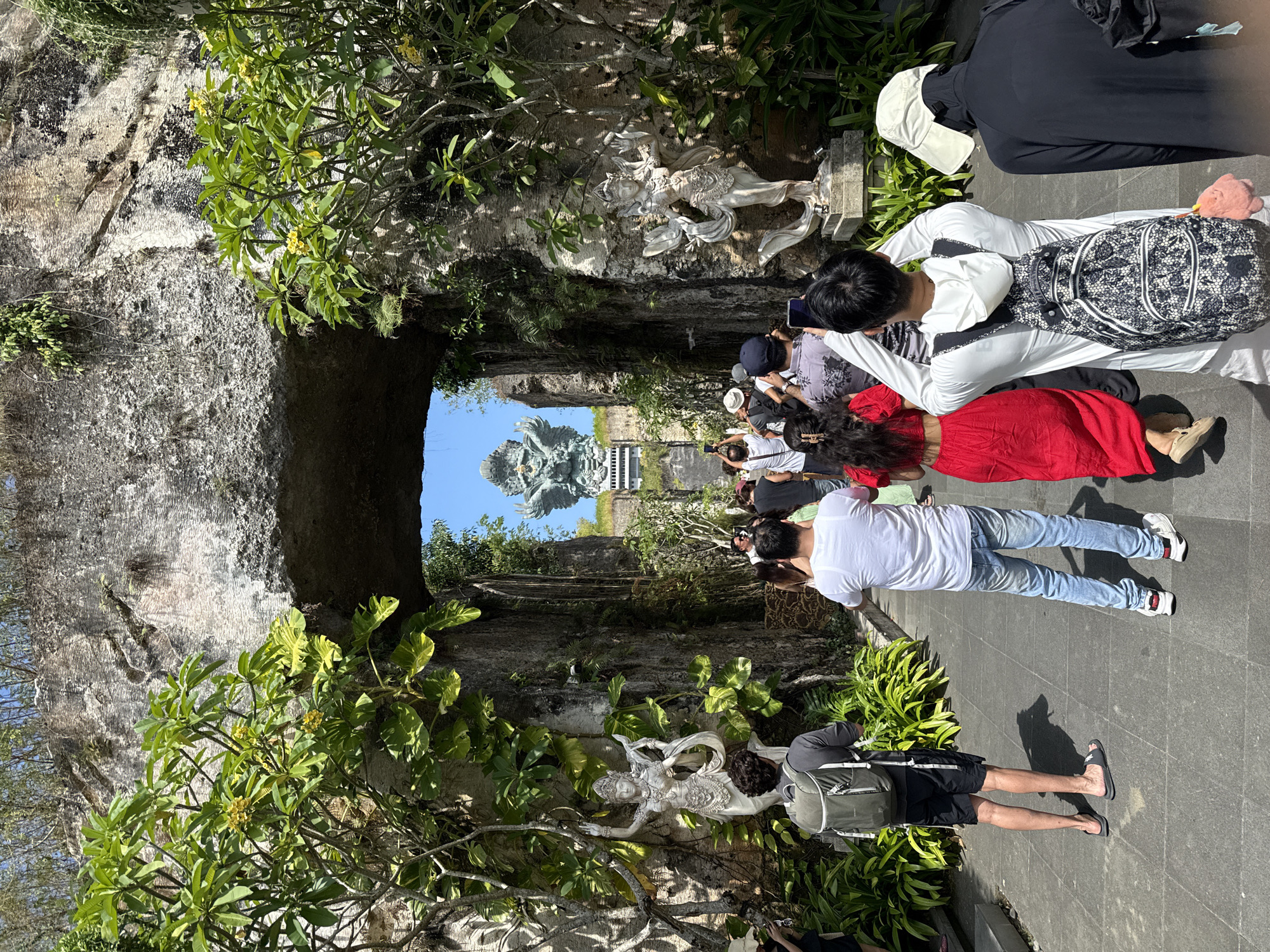 A horizontal eye-level shot depicts a group of people looking through a stone structure at the Garuda Wisnu Kencana statue in Kuta Selatan, Indonesia. The foreground is framed by a weathered stone structure with lush greenery and decorative statues clinging to its sides. A group of people, varying in dress, are positioned along a paved path, gazing towards the statue. Some individuals are holding up phones, presumably taking photographs. The background features the prominent Garuda Wisnu Kencana statue, set against a clear blue sky. The statue stands tall and detailed.