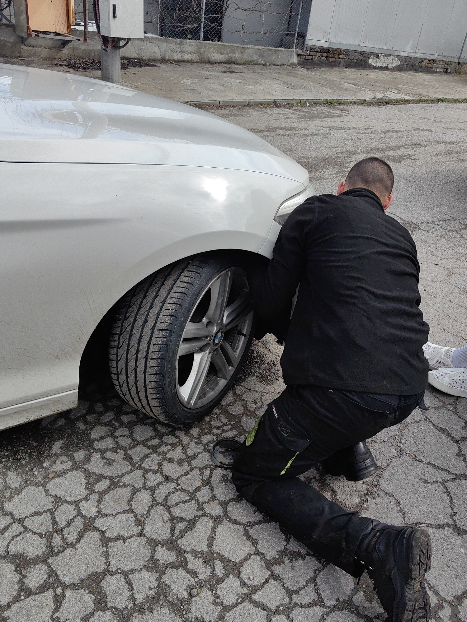 A man is kneeling near the front driver's side tire of a white BMW car. The man is wearing a black jacket, black pants with green accents, and black boots. He appears to be examining the tire or wheel well. The car is parked on a cracked asphalt surface. In the background, there is a chain-link fence and a white building. A pair of white sneakers are visible near the man. The location is possibly in Sofia, Bulgaria.