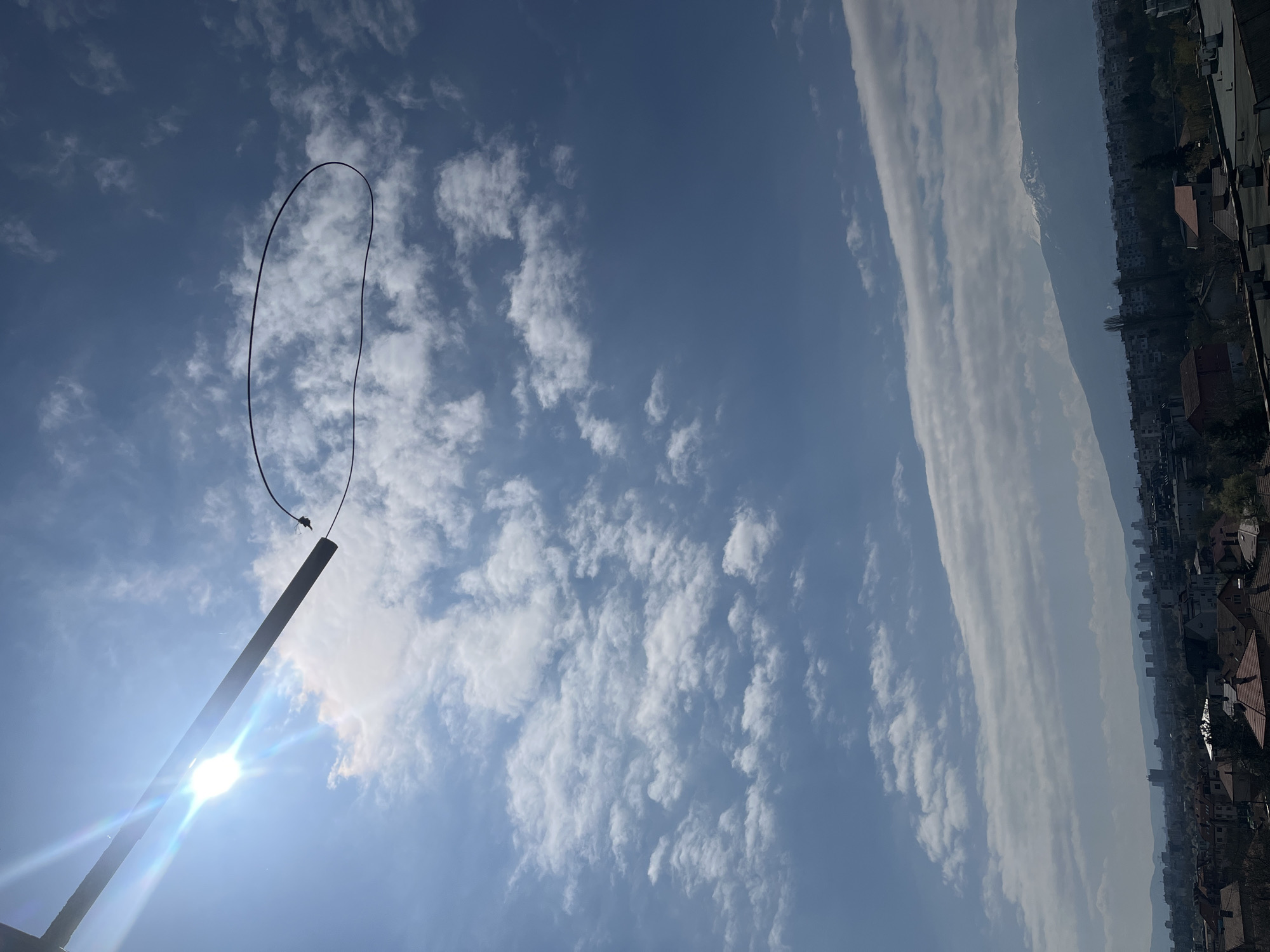 This image presents a bright daytime sky in Sofia, Bulgaria. The sky is predominantly blue, scattered with puffy white clouds. On the right side of the frame, a long, thin band of clouds stretches horizontally across the horizon, partially obscuring buildings. A tall, dark-colored pole is visible at the lower left of the frame, extending upwards into the sky with a wire or cable loop attached to its top. Sunlight appears to be directly behind the pole, resulting in significant lens flare. The photograph appears to be taken from a high vantage point.