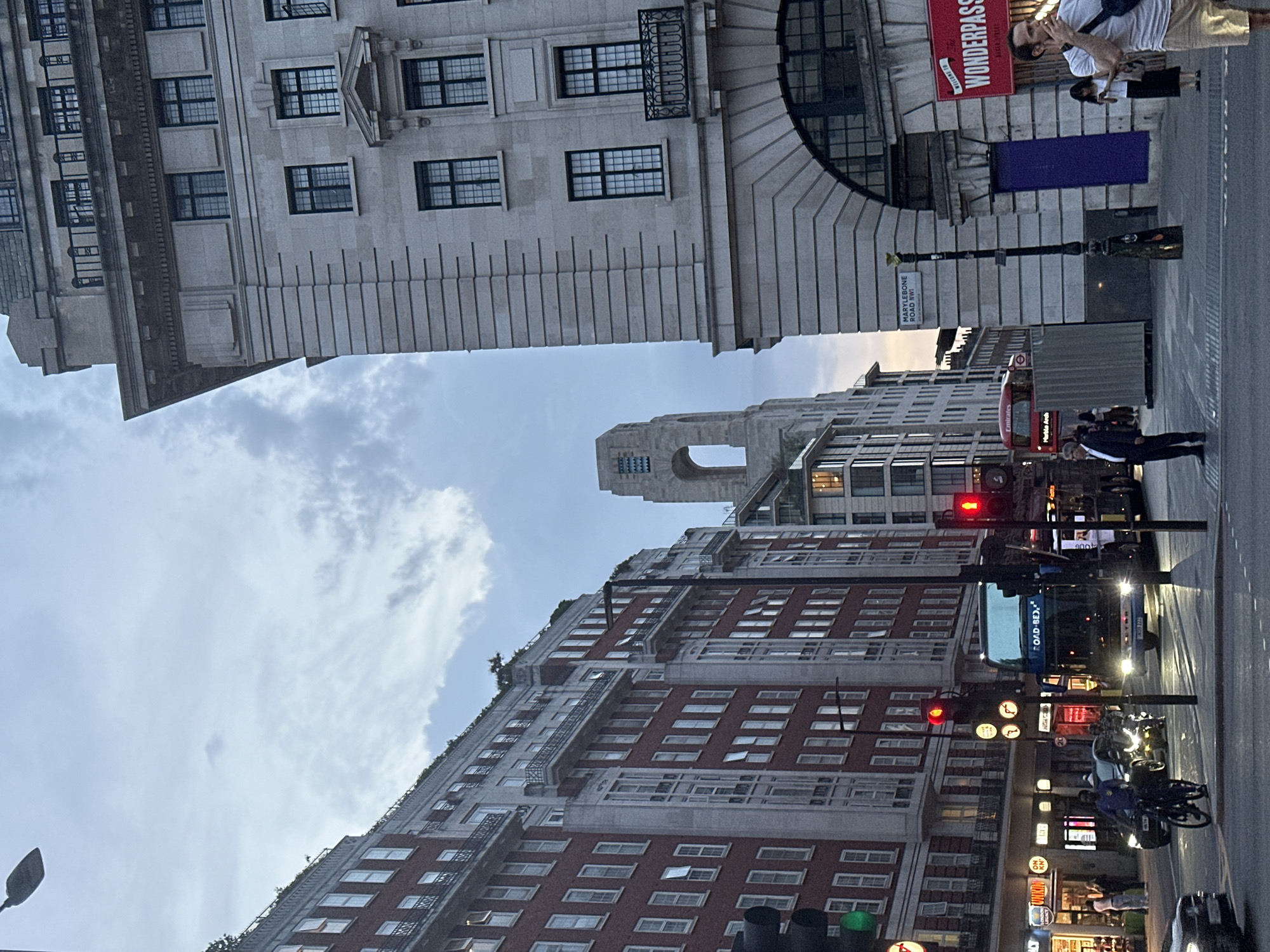 The image shows a street scene in London, United Kingdom. The view is framed by buildings on either side, revealing a glimpse of the sky. On the left, a tall building with numerous windows rises. On the right, a building with arched architecture is visible, with a sign reading "WONDERPASS" and a street sign indicating "MARLEYBONE ROAD BAKER ST."
Pedestrians are present on the street, some appearing to be interacting and others walking along the sidewalk. Vehicles, including a double-decker bus, are on the road. Traffic lights are visible, indicating red. Overall, the scene depicts a typical urban environment with architectural landmarks, pedestrians, and vehicular traffic.