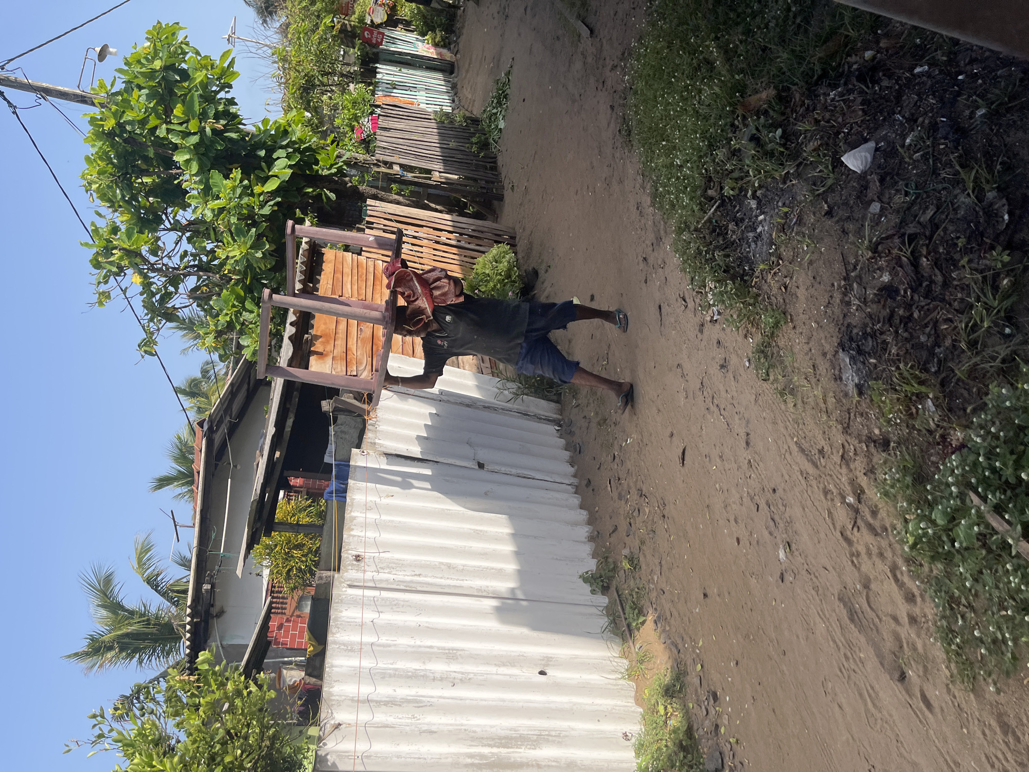A man is carrying a large, pink plastic chair over his head and shoulders, on a dusty dirt road in a semi-urban area. He is wearing a dark green polo shirt and blue shorts. The chair is wrapped in a red and white cloth at the top. To his left is a corrugated metal fence, and behind that, buildings with corrugated roofs and palm trees are visible. To his right, there is a wooden fence and more greenery. The sky is clear and blue. The scene appears to be in Moratuwa, Sri Lanka, a coastal city known for its furniture manufacturing.