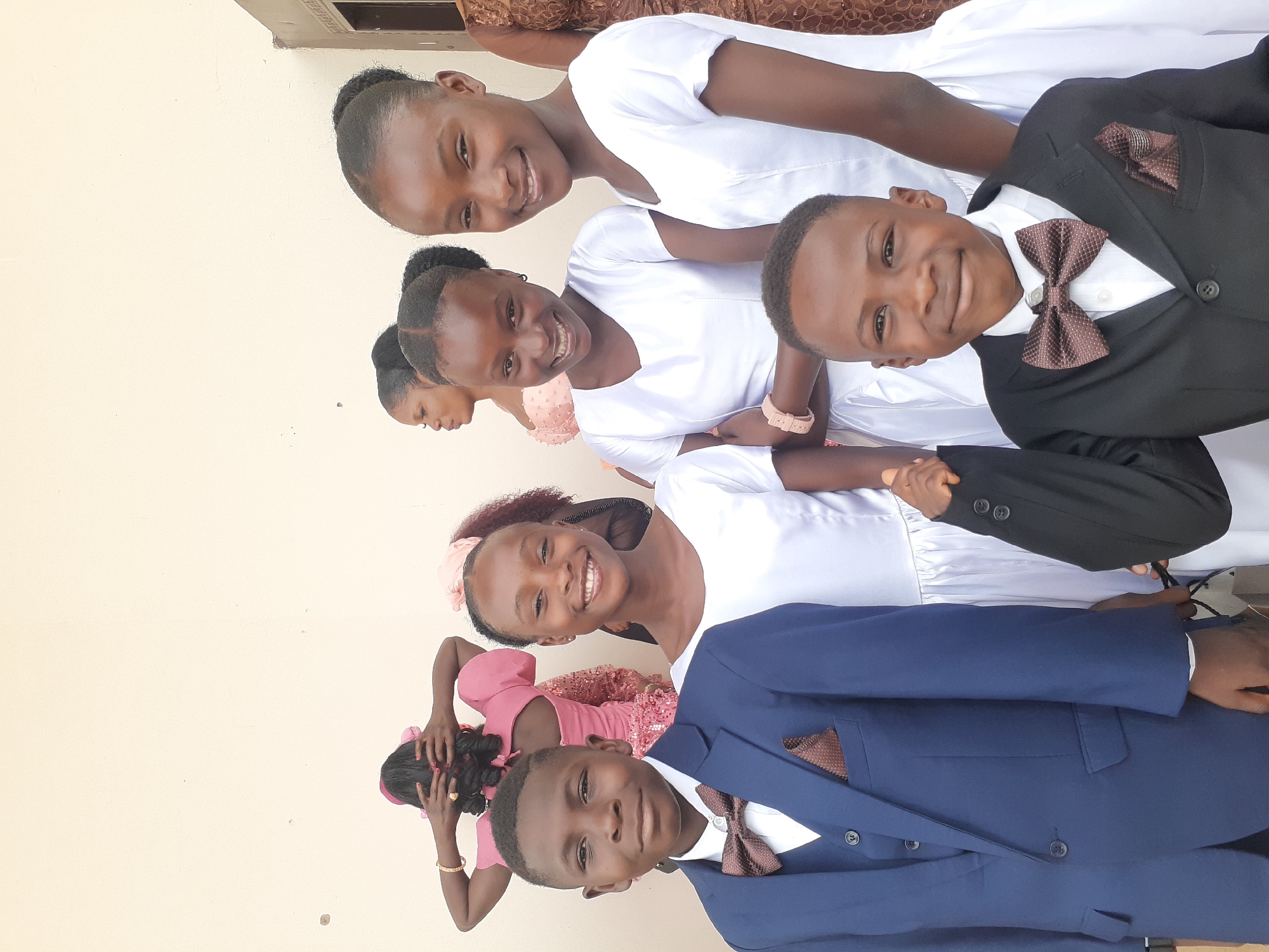 A group of children are posing outdoors, likely for a formal event. There are three girls wearing white dresses and two boys wearing suits with bow ties. One boy wears a navy suit while the other boy is wearing a black suit. Two girls are wearing pink dresses. The background is a light-colored wall, possibly of a building in Barakin Lerawi, Nigeria. The image is taken in portrait orientation.