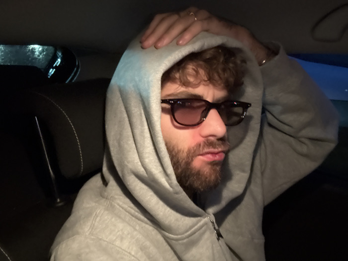 An adult male is depicted seated inside a vehicle, likely in the rear passenger compartment. He has curly, reddish-brown hair visible beneath a grey hooded sweatshirt with the hood drawn over his head. He wears dark-framed sunglasses with reddish-tinted lenses, and a full reddish-brown beard and mustache. His right hand is raised, with fingers resting on the top of his head; a ring is visible on the ring finger and a silver-colored band on the wrist. His mouth is slightly open with lips pursed. The vehicle interior features dark-colored seating with white stitching visible on a headrest to the left of the subject. The background to the left reveals a blurred window, showing indistinct lights or reflections suggesting an external environment that is dark, possibly with rain on the glass. The overall lighting is dim. No specific identifying features or landmarks related to the city of Givors, France, are discernible within the image.