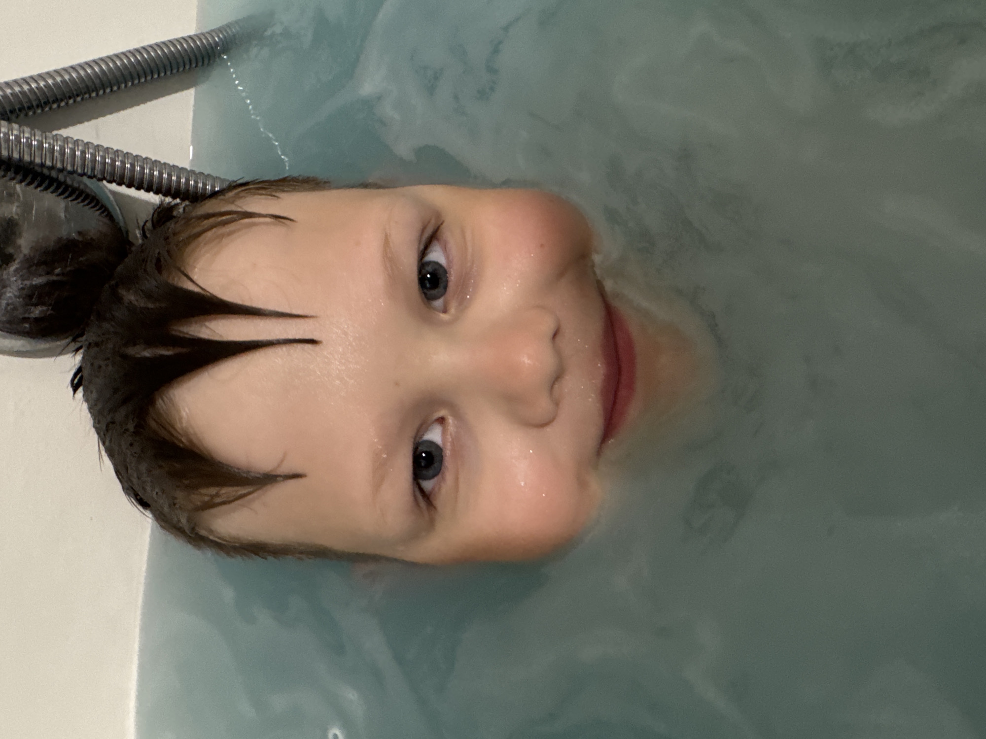 A young child is shown partially submerged in a bathtub filled with light blue water. The child's head is tilted back, and their face is looking upwards towards the camera. Their eyes are open and they appear to be smiling. Their hair is dark and wet, and some strands are sticking to their forehead. The child's nose is slightly shiny, suggesting it's wet. The bathtub appears to be made of a white material. A portion of a chrome shower hose is visible in the upper left corner of the image. The water is calm, with some ripples and swirls visible. The lighting suggests it is likely indoors, and the time of day cannot be determined. There is no visible text in the image.
