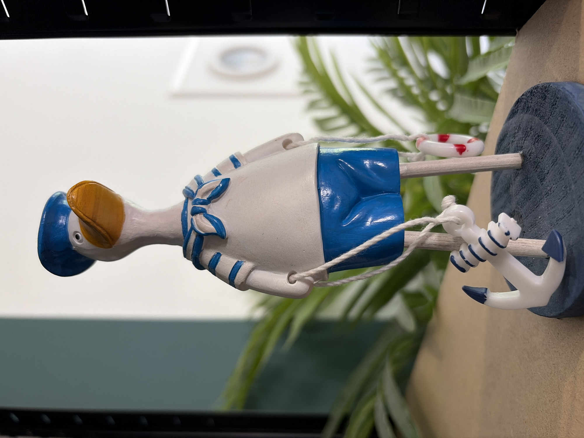 A ceramic figurine of a duck dressed as a sailor stands on a wooden base. The duck is wearing a blue sailor hat, a white shirt with blue trim, and blue shorts. It is holding white ropes in its hands, which are attached to a small anchor and a life preserver. The figurine is positioned on a light brown surface with a blurred green plant in the background. The background also shows a white wall with a circular object and a black vertical object on the right side. The location is not discernible from the image.