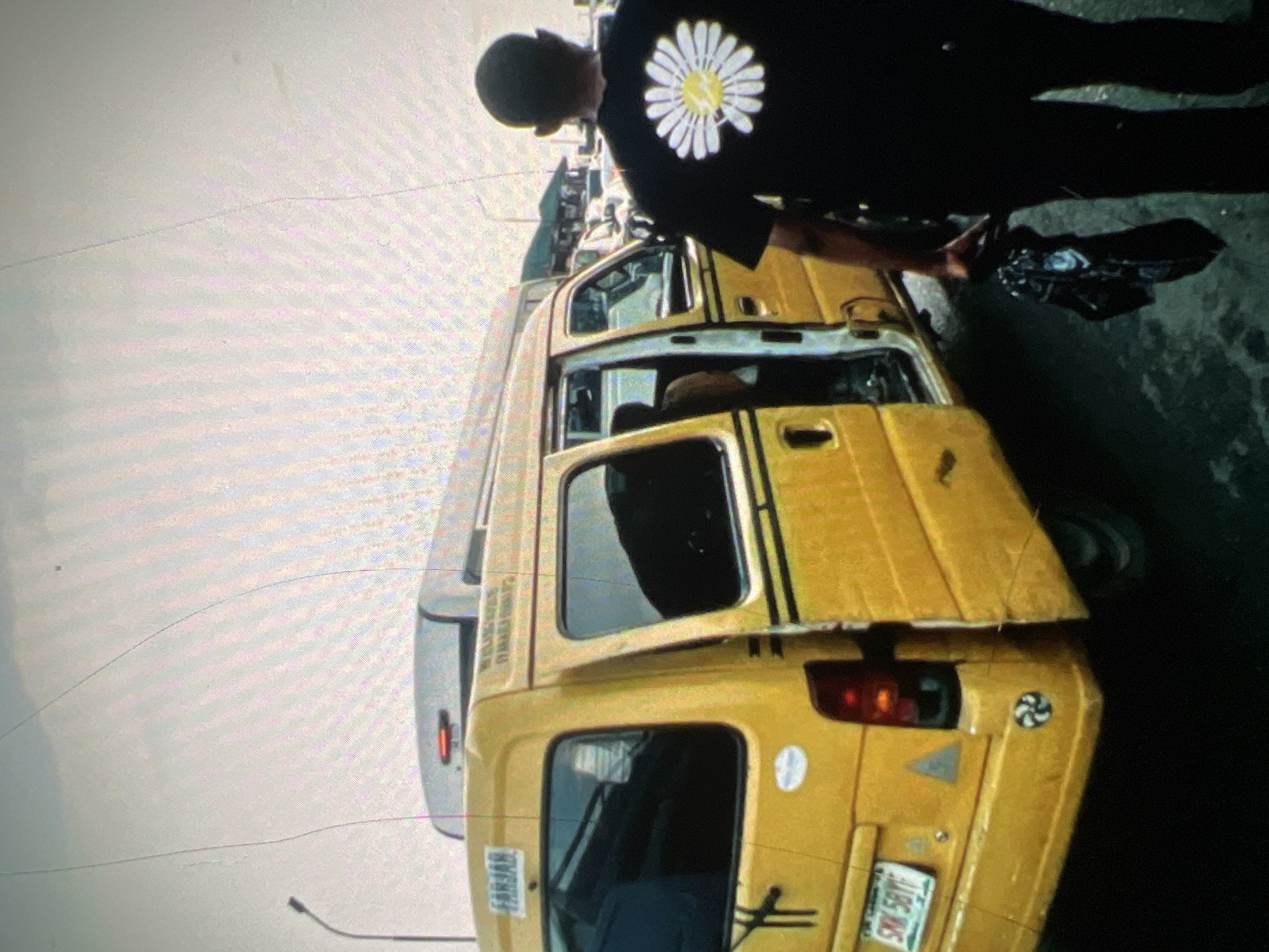 In Lagos, Nigeria, a yellow minibus, commonly known as a danfo, is parked on a street. The minibus has its rear door open and appears to be picking up or dropping off passengers. A man, seen from behind, stands next to the open door, holding a black bag. He is wearing a black t-shirt with a white daisy graphic on the back. The street is wet, suggesting recent rain or it being a drainage area. Other vehicles and buildings are visible in the background, indicating an urban setting. The sky is overcast.
