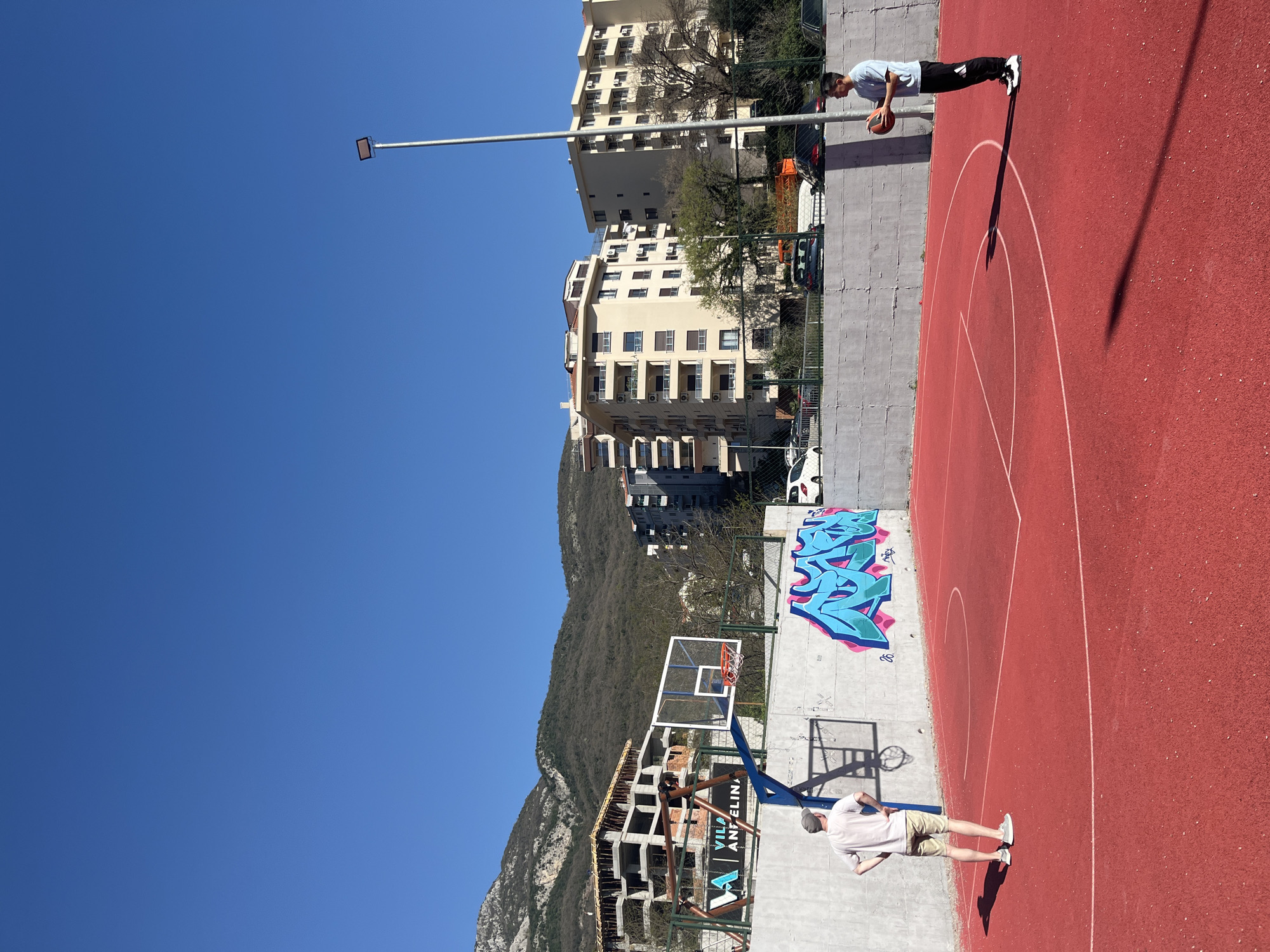 The image captures an outdoor basketball court in Rafailovići, Montenegro, under a clear, bright blue sky. The court is red and marked with white lines delineating the basketball zones. Two individuals are present: One stands near the top of the image, dressed in a light-colored shirt and dark pants, holding a basketball. The other person stands near the lower right corner of the image wearing a light-colored t-shirt and khaki shorts. Graffiti adorns a section of the court's wall near the basketball hoop. In the background, multi-story buildings and green trees are visible, with the slopes of a forested mountain rising further back. A light pole extends upwards to the left of the buildings. The angle of the photograph suggests it may have been taken from an elevated position.