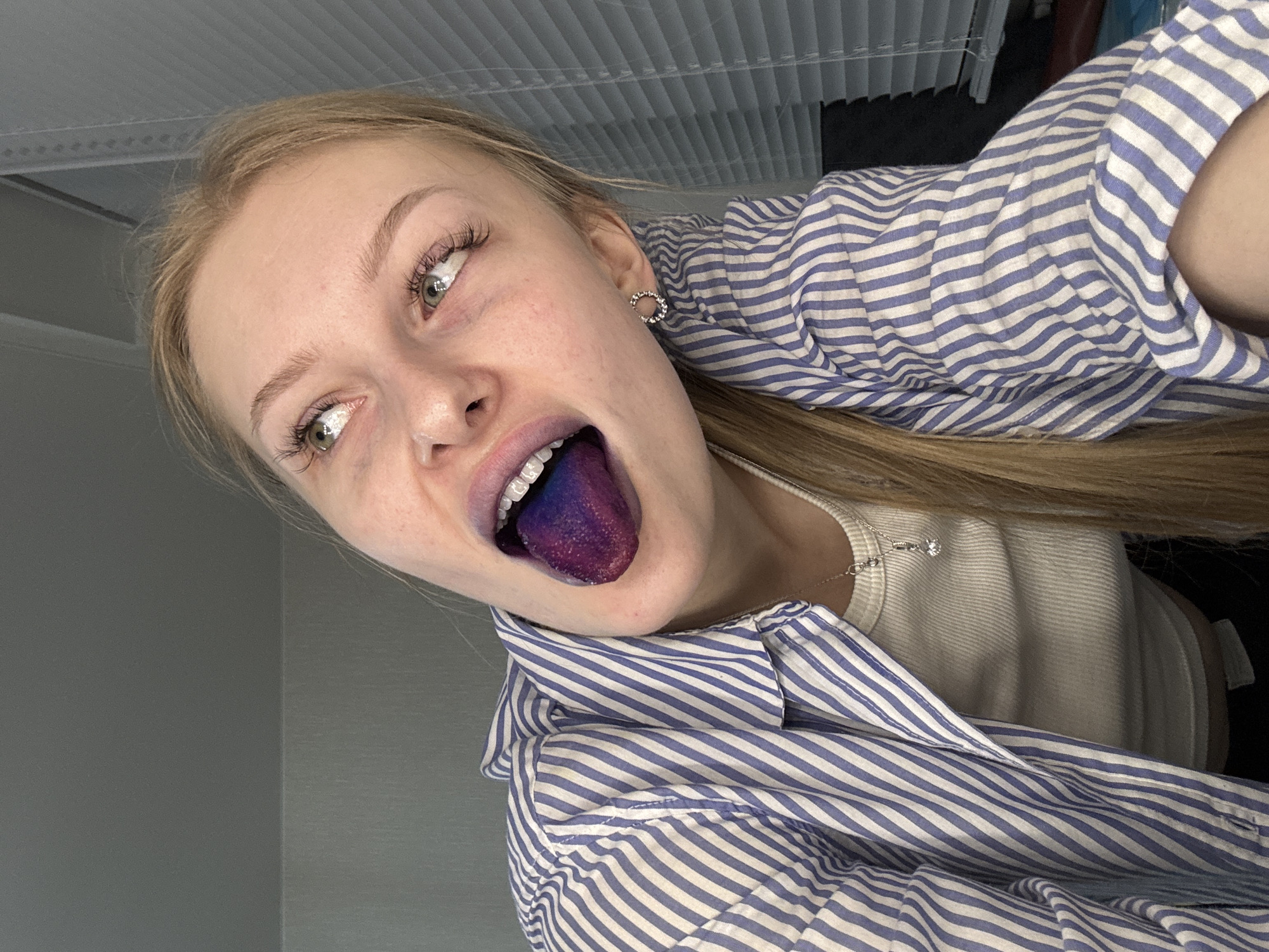 A close-up, angled portrait features a young woman with light blonde hair, fair skin, and green/hazel eyes, looking upwards and to the right. Her mouth is open in a playful expression, revealing a tongue dyed a vivid purple and blue hue. She is wearing a blue and white striped collared shirt, unbuttoned to show a light-colored ribbed top underneath. A delicate silver chain with a small pendant adorns her neck, and a silver hoop earring with embedded stones is visible in her right ear. The indoor setting, located in Perm, Russia, includes plain light gray walls and a ceiling with visible horizontal blinds or a textured surface. The lighting is soft and even. The image conveys a lighthearted and expressive mood. No visible text is present.