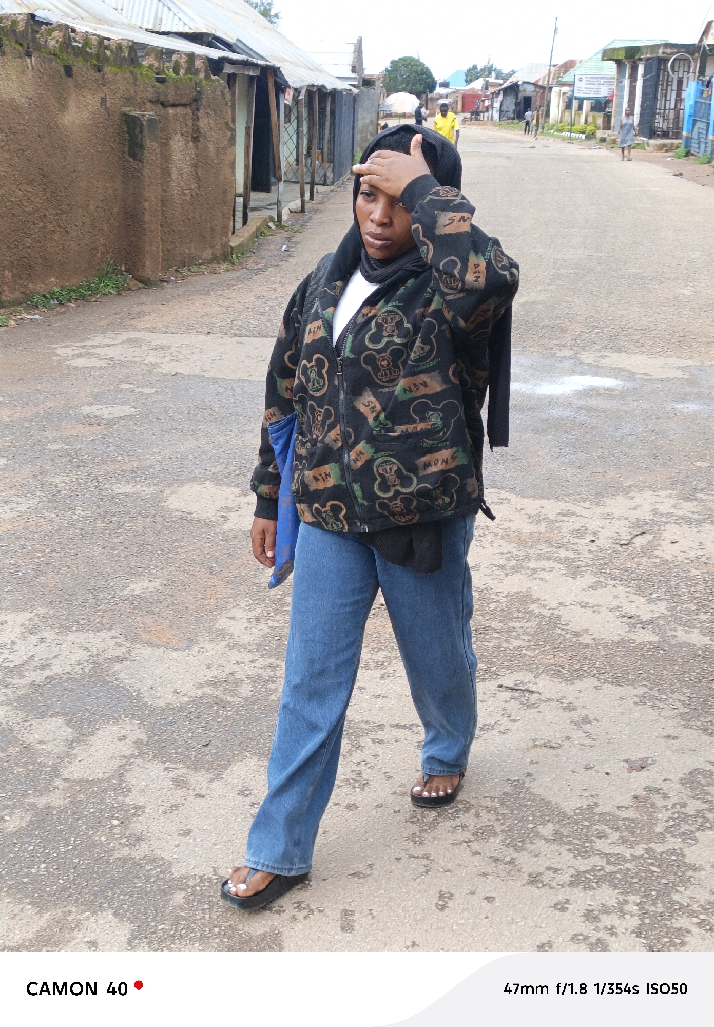 A young woman, wearing a black hijab and a patterned hooded jacket over a white t-shirt, walks on an asphalt road in Jos, Nigeria. She has her right hand on her forehead as if shielding her eyes from the sun. She is wearing blue jeans and black sandals. The road is lined with buildings that appear to be made of mud and corrugated metal. In the background, other people are visible, as well as some trees and a cloudy sky. A blue bag is slung over her left arm.