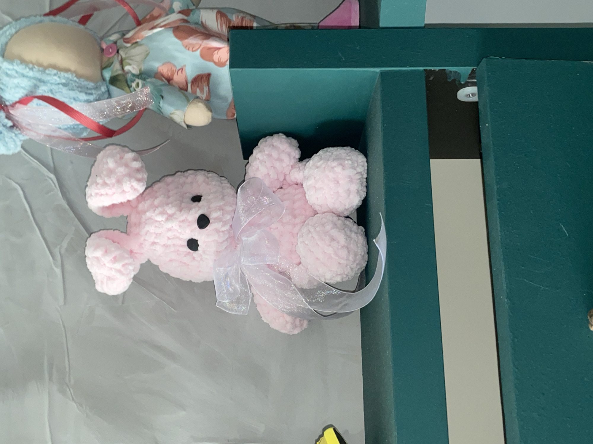 An overhead shot features a fluffy pink crocheted bunny with black eyes and a white ribbon tied around its neck, sitting on a teal shelf. To the right of the bunny, a doll with blue hair and a floral-patterned dress is partially visible. The background is a textured gray wall. The shelf is dark teal and sits in front of a wall. The image appears to be taken indoors.