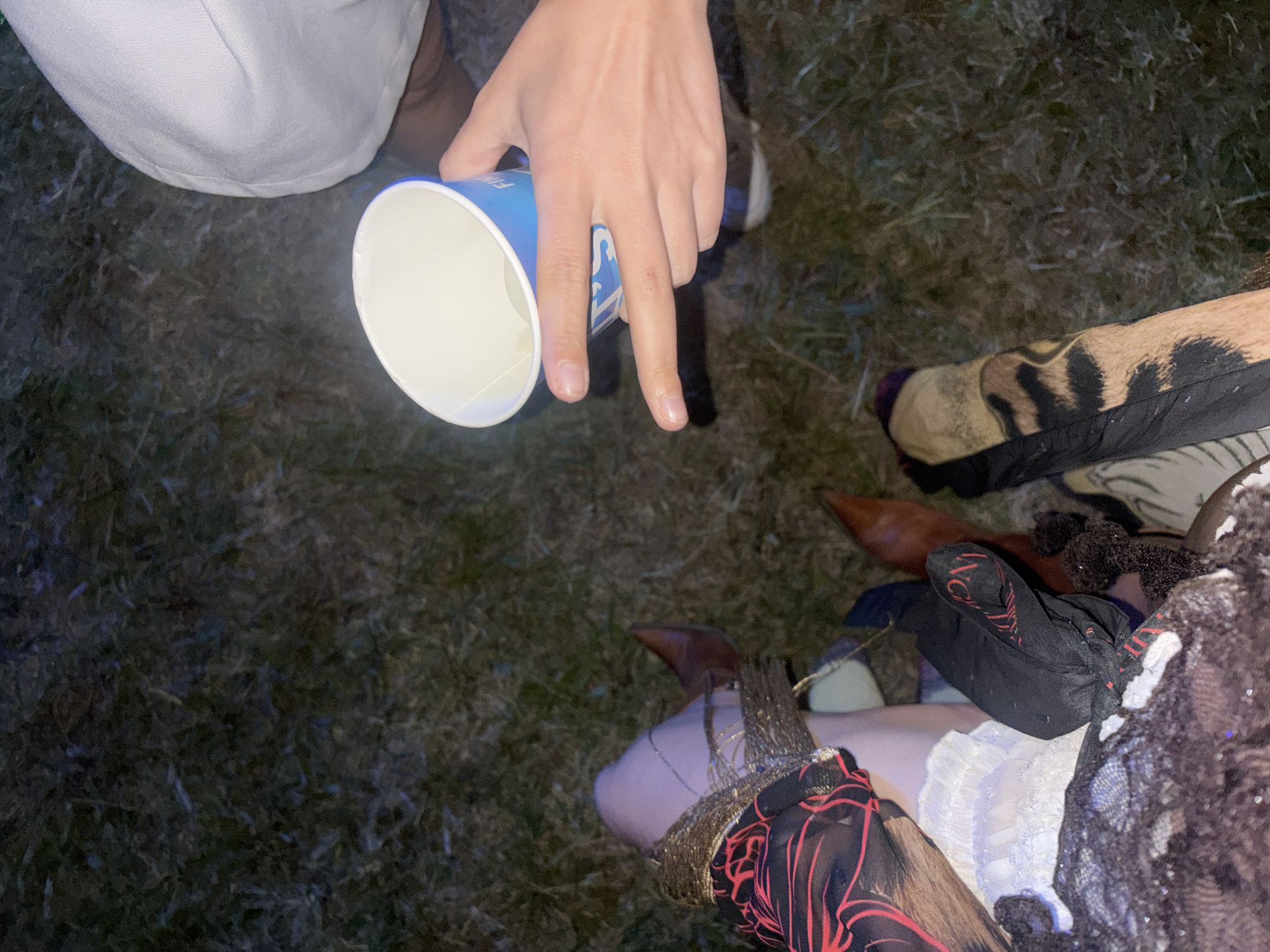 The image shows a close-up view of several people's legs and hands, possibly gathered outdoors on a grassy area. One person's hand is holding a blue and white disposable cup, with a slight liquid level visible inside. The legs are adorned with various items, including brown boots, black gloves with red accents, sheer patterned fabric with red designs, a white lace-trimmed garment, and a garment with a tiger-like print. The lighting suggests it might be evening or night. There is no discernible information in the image to indicate a specific location within the city of Sofia, Bulgaria.
