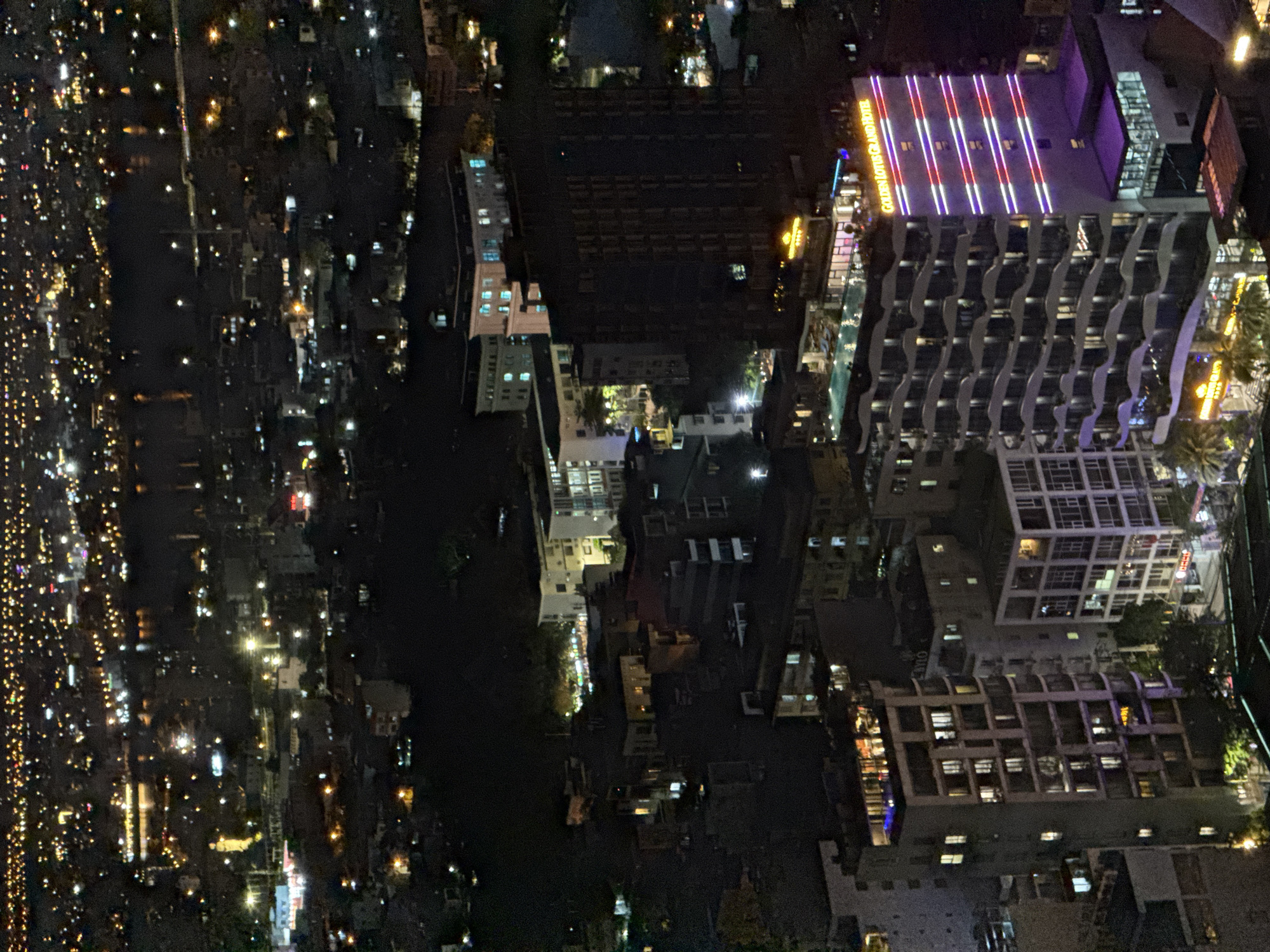 This is a nighttime aerial view of the city of Đà Nẵng, Vietnam, featuring a cityscape illuminated by numerous lights. In the foreground, the "Golden Lotus Grand Hotel" is prominently visible, distinguished by its lit-up facade displaying horizontal stripes of purple and pink lights. The hotel has a tiered, wavy architectural design. Adjacent to it, another building with a swimming pool and lounge chairs on its rooftop is visible. Other buildings, some with lit windows and signs, populate the surrounding urban landscape, extending into the distance where a body of water is discernable, reflecting city lights. The overall scene depicts a bustling urban environment at night.
