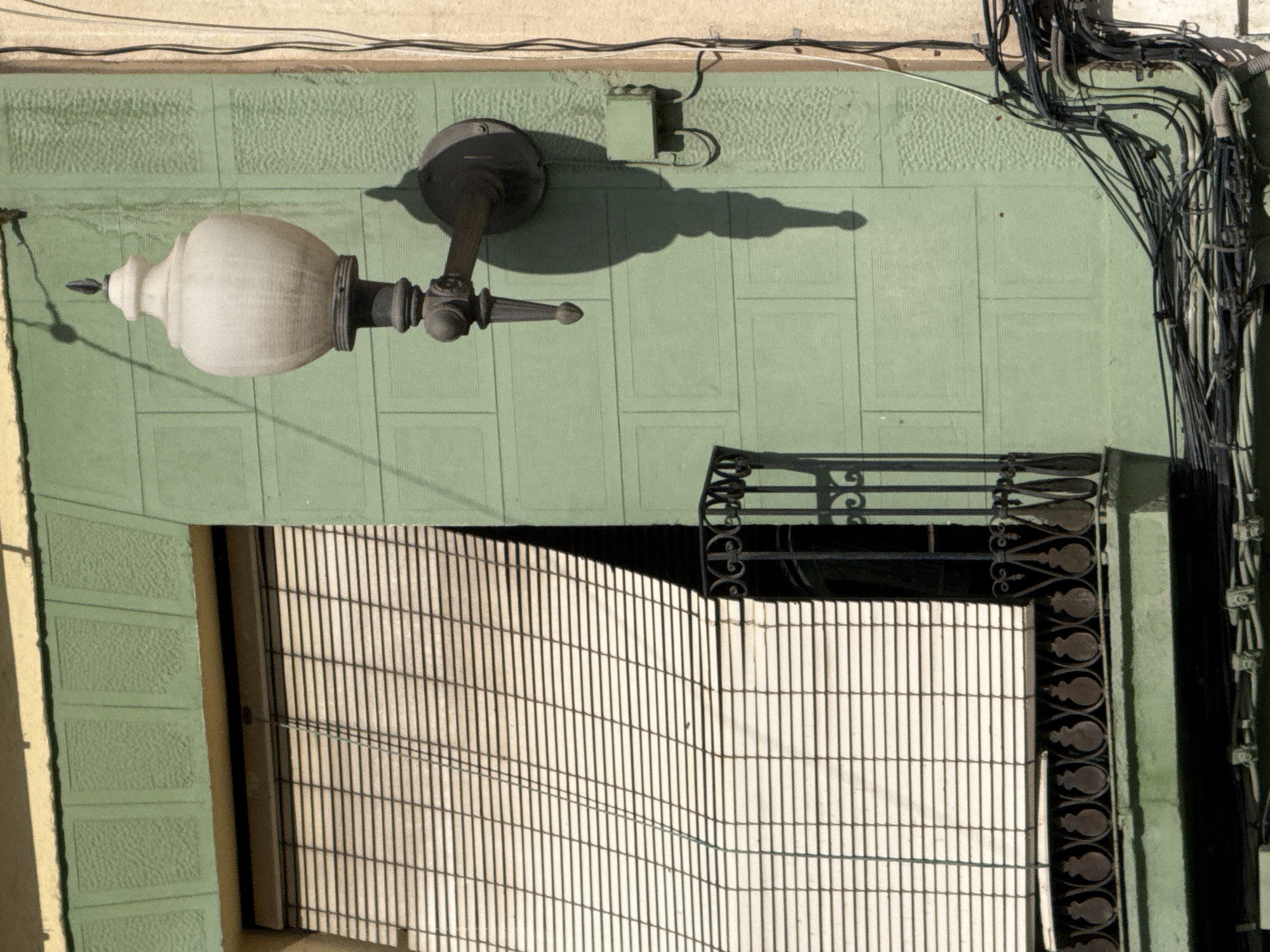 The image displays a detailed, vertical close-up of a building facade in the city of Unknown, Unknown, under bright daylight conditions. The main section of the facade is painted a muted green, featuring recessed rectangular panels, some with a smooth finish and others with a distinct pebbled texture. A narrow, horizontal strip of yellow wall is visible at the very top, and a vertical strip of lighter tan-colored wall is present on the far right edge of the frame.
On the left side of the facade, a window is completely covered by an exterior slatted blind of an off-white or light beige color. The blind is partially rolled down and appears slightly angled, casting subtle horizontal shadows across its slats, indicating a strong light source from the upper right. Below the window is a small balcony equipped with a dark wrought iron railing, characterized by a repeating pattern of stylized, inverted pear shapes or scrolls.
To the right of the window and balcony, a vintage-style wall-mounted light fixture is prominently featured. It consists of a dark-colored metal arm, possibly cast iron, supporting a large, opaque, off-white or cream-colored globe-shaped lamp shade. The fixture is topped with a decorative finial and has a pointed element extending from the bottom of its arm. The intense sunlight casts a long, distinct, dark shadow of the entire light fixture directly below it on the green wall, extending downwards.
The lower right portion of the green wall is densely covered with numerous black electrical wires and cables. These cables are bundled, routed along the wall, and appear somewhat tangled, indicative of utility installations. Some conduits and junction box elements are visible within this cluster of wiring. No people or animals are present in the scene.