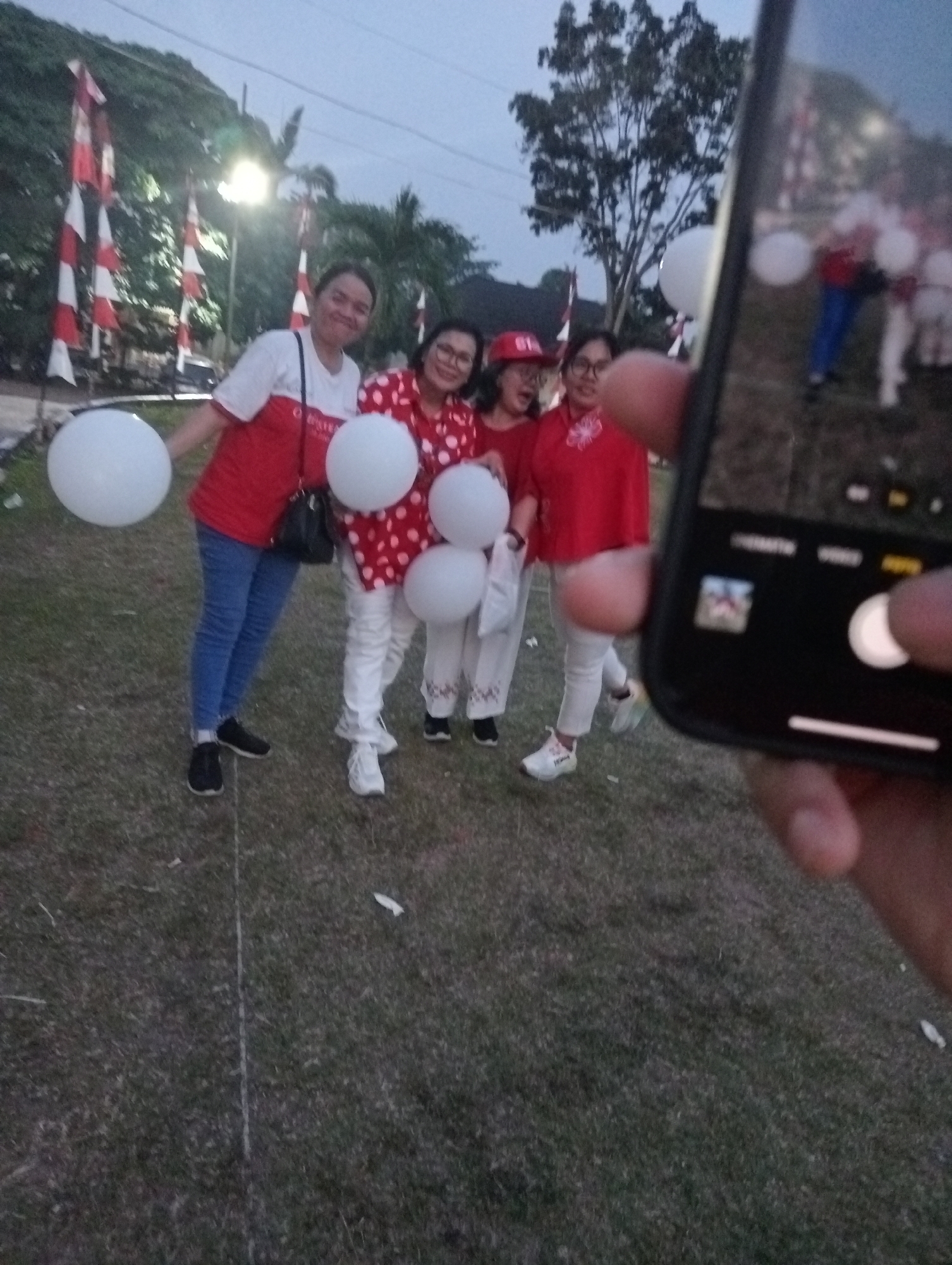 In Jambi, Indonesia, four women are posing for a photograph in an outdoor grassy area with trees and flags in the background. The woman on the far left is wearing a white shirt with red accents and blue jeans, holding a white balloon. Next to her, a woman in a red polka dot shirt and white pants smiles, also holding a white balloon. The third woman, wearing a red shirt and white pants, holds a white balloon, and the woman on the far right is wearing a red shirt and white pants. The scene appears to be a casual gathering, possibly a celebration, given the balloons and the festive attire. The lighting suggests it is either early morning or late evening. The photograph is being taken by someone holding up a smartphone, the screen of which is visible in the foreground, partially obscuring the group.