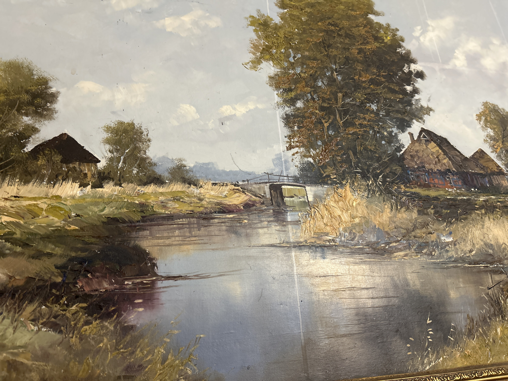 The image is an oil painting depicting a rural landscape. A winding river dominates the foreground, reflecting the sky. On the left bank, there is a thatched-roof building nestled among trees. A small bridge spans the river further back. On the right bank, another cluster of thatched-roof buildings is partially obscured by a large tree. The sky is overcast with scattered clouds. The location appears to be a rural area, perhaps in the countryside.