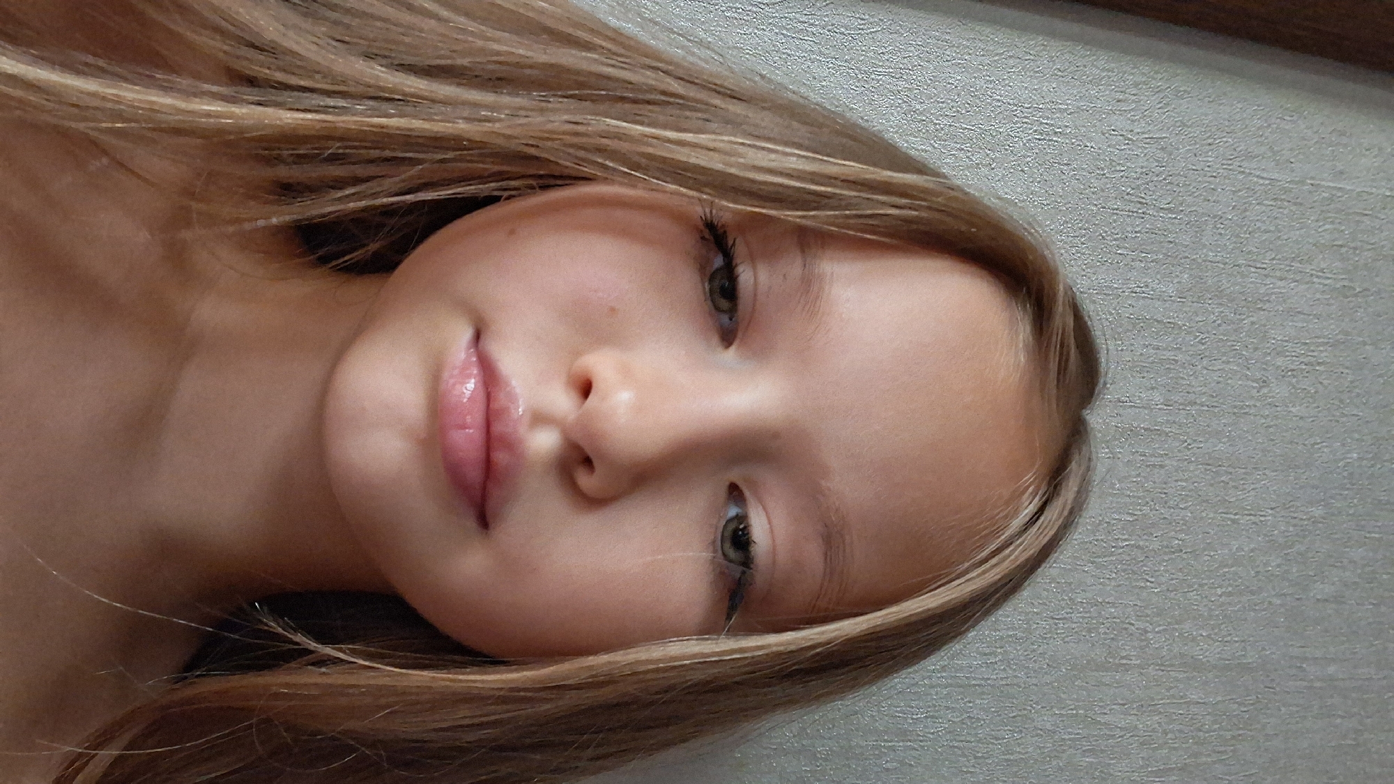 The media file is a close-up portrait of a young girl with long, blonde hair. She is lying down, with her head tilted to the side, and her hair is spread around her. Her eyes are open, and she appears to be looking upwards. The lighting suggests an indoor setting, likely daytime given the natural light illuminating her face. Her expression is neutral or slightly contemplative. The background is a textured, light-colored wall. The image is oriented vertically, making it a portrait orientation. No text is visible in the image. The location context is Semey, Kazakhstan.