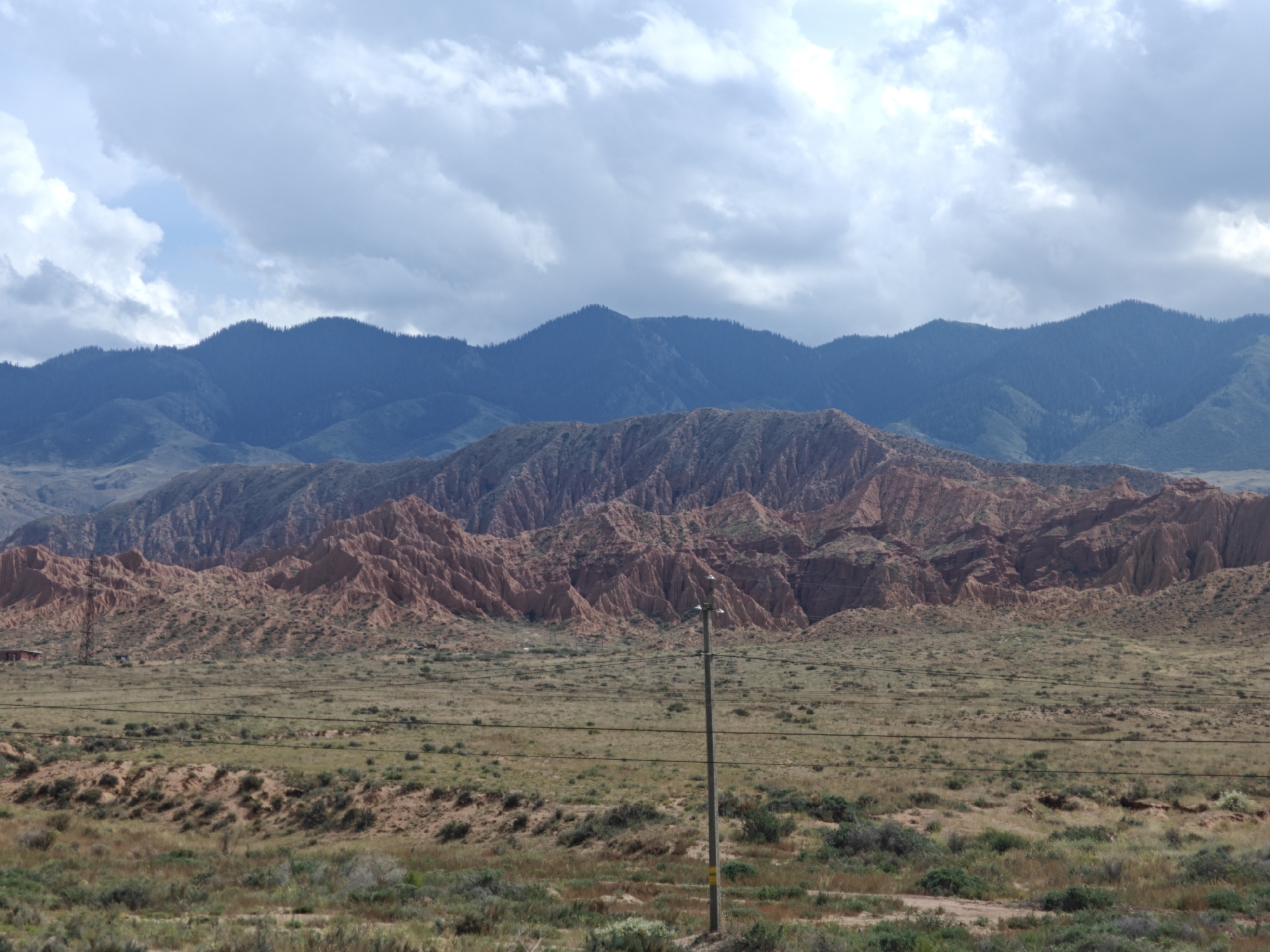 The image depicts a vast, arid landscape under a cloudy sky. In the foreground, a flat, grassy plain stretches towards a range of undulating hills and mountains. Several power lines crisscross the scene, supported by a wooden pole in the middle ground. In the far distance, a range of darker, more densely vegetated mountains forms the horizon. The terrain appears rugged and eroded, with the hills in the middle ground showcasing distinct layers of reddish-brown rock. The sky is filled with heavy, gray clouds, with patches of lighter blue sky visible. The overall impression is one of a remote, windswept natural environment. The presence of power lines suggests human infrastructure, but the scene is otherwise sparsely populated. The location is identified as Kaji-Say, Kyrgyzstan.