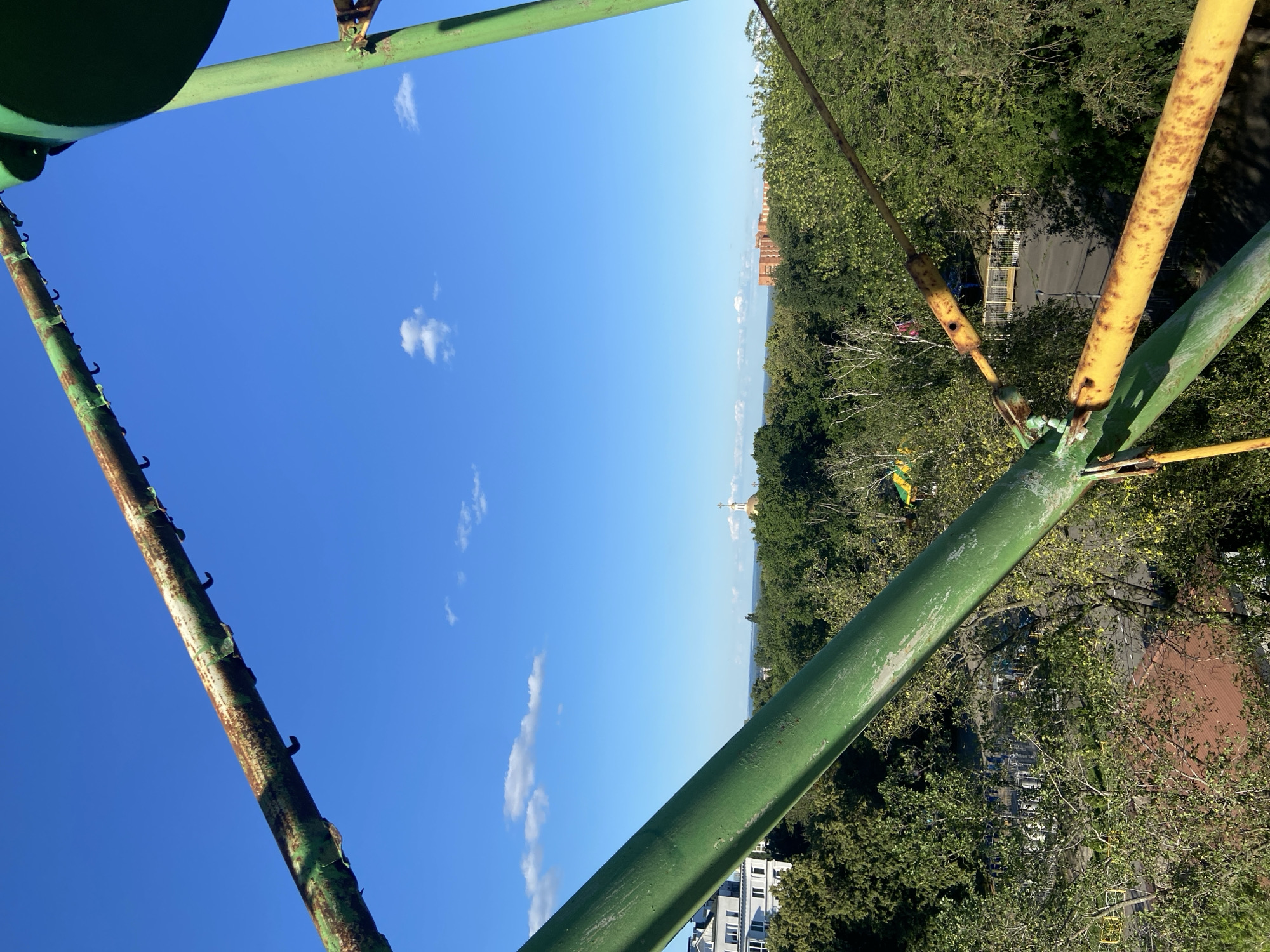 The image is an upward-looking shot from a metal structure, possibly a Ferris wheel or observation tower, in Nyzhni Mlyny, Ukraine. The primary subjects are the large, green and yellow metal beams of the structure itself, which dominate the foreground and are weathered with rust. These beams create a framing effect, guiding the viewer's eye towards the expansive blue sky dotted with a few white clouds.
Beyond the structure, the scene opens up to a panoramic view of a densely wooded area interspersed with buildings. A few notable structures are visible in the distance: a brick building with multiple windows, and further on, a church with a golden dome. The foliage is lush and green, suggesting a warm weather season. A road can be seen winding through the trees, with hints of yellow playground equipment.
The time of day appears to be late morning or early afternoon, judging by the bright sunlight and the clear blue sky. The weather seems pleasant and sunny, ideal for outdoor activities. The perspective from a height suggests a location offering a vantage point over the town and its surroundings. There is no visible text in the image. The overall impression is one of scenic beauty and a sense of elevation, offering a glimpse of the Ukrainian landscape.