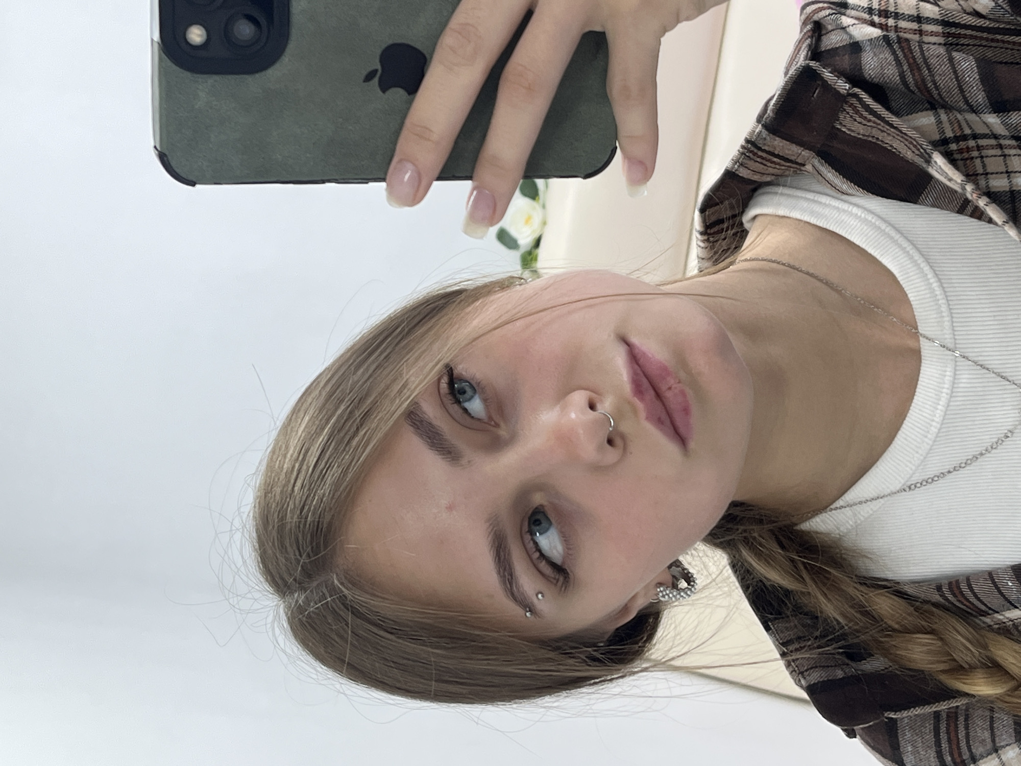 A young woman is taking a selfie. She is wearing a white tank top and a brown and white plaid shirt. She has blonde hair and blue eyes. She has a nose ring and an eyebrow piercing. She is holding a smartphone with a green case. The background is white and nondescript. The location is unlikely to be determined based on the image content.
