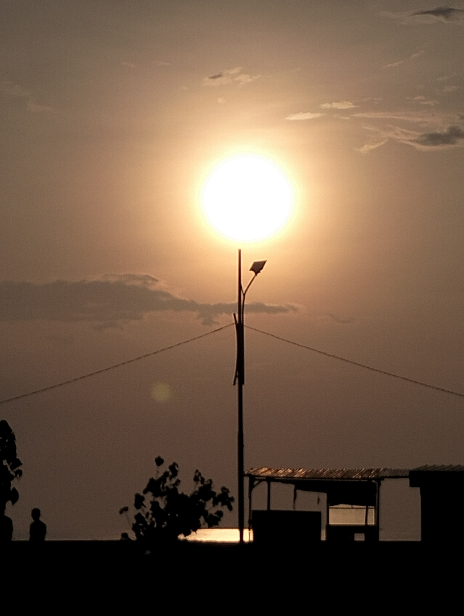 The image captures a serene, back-lit scene during what appears to be either sunrise or sunset over a body of water in Lhokseumawe, Indonesia. The sky dominates the upper two-thirds of the frame, displaying warm, hazy hues of orange, brown, and light yellow, with sparse, wispy clouds visible against the diffused glow.
A brilliant, intense white sun, partially softened by atmospheric haze, is centrally positioned, acting as the primary light source. Directly beneath the sun stands a tall, dark silhouetted streetlight featuring a distinct light fixture and a solar panel at its top. Two thin, horizontal wires extend from the pole, traversing the mid-ground.
The lower third of the image consists of dark silhouettes. On the left, a solitary human figure is visible, standing by what appears to be the edge of the water or land. Adjacent to the person, and slightly to the right, are silhouetted clusters of bushy foliage. Further to the right, several structures are silhouetted against the hazy light, including a shelter with a corrugated roof and visible openings, suggesting a simple building or stall. A faint glimmer of light is reflected on the implied water surface just above these foreground elements.
The overall mood is peaceful and contemplative, with the strong backlighting creating dramatic contrast and emphasizing the outlines of all foreground subjects. No visible text is present in the image.