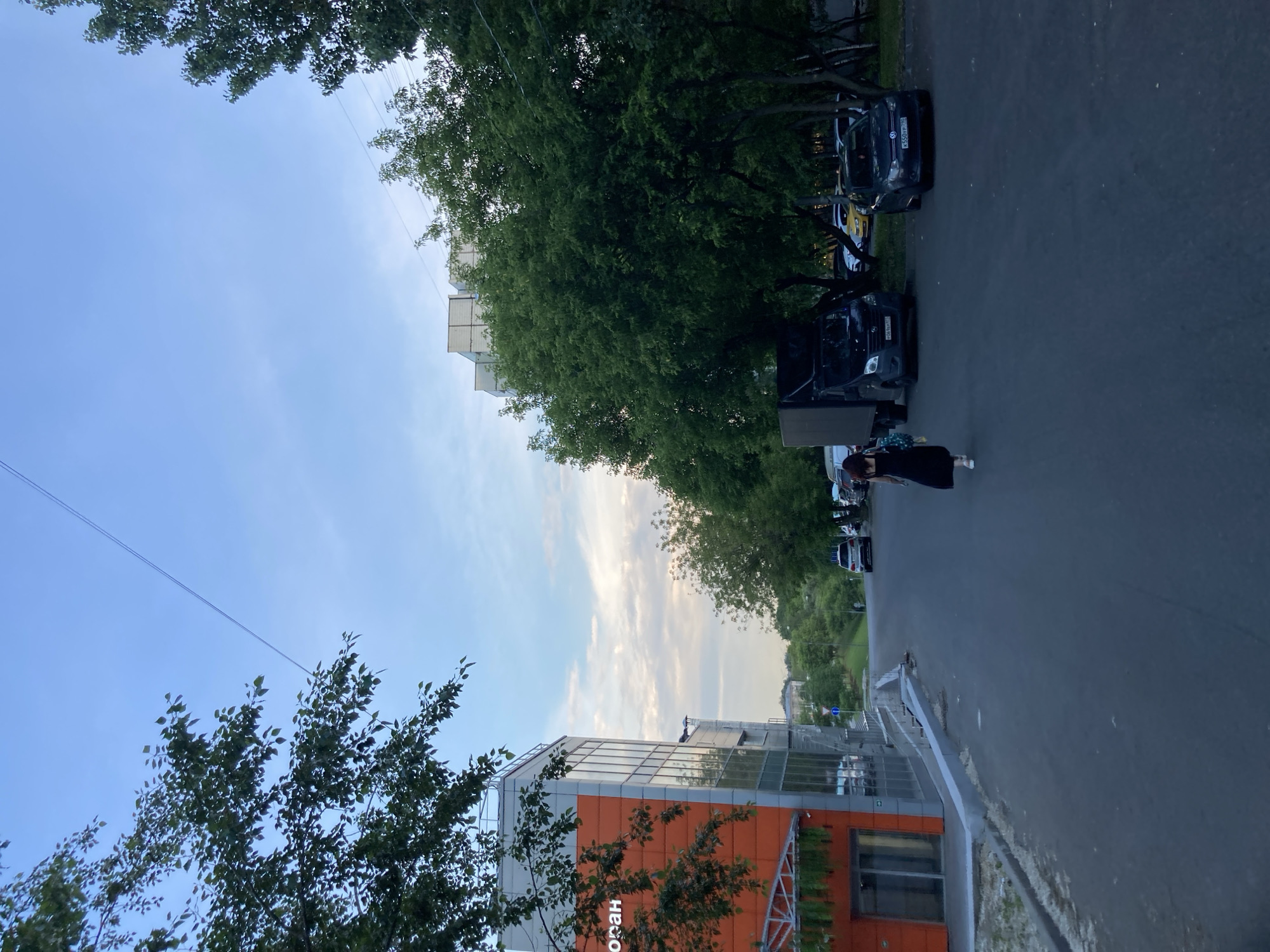 A woman in a black dress walks away from the camera on a paved street. To her left is an orange and grey building with large windows, and to her right are several parked cars and trees. A black truck is parked in the middle of the street. The sky is partly cloudy and blue. The scene is set in a city, likely Moscow, Russia, given the architecture and vehicle license plates.