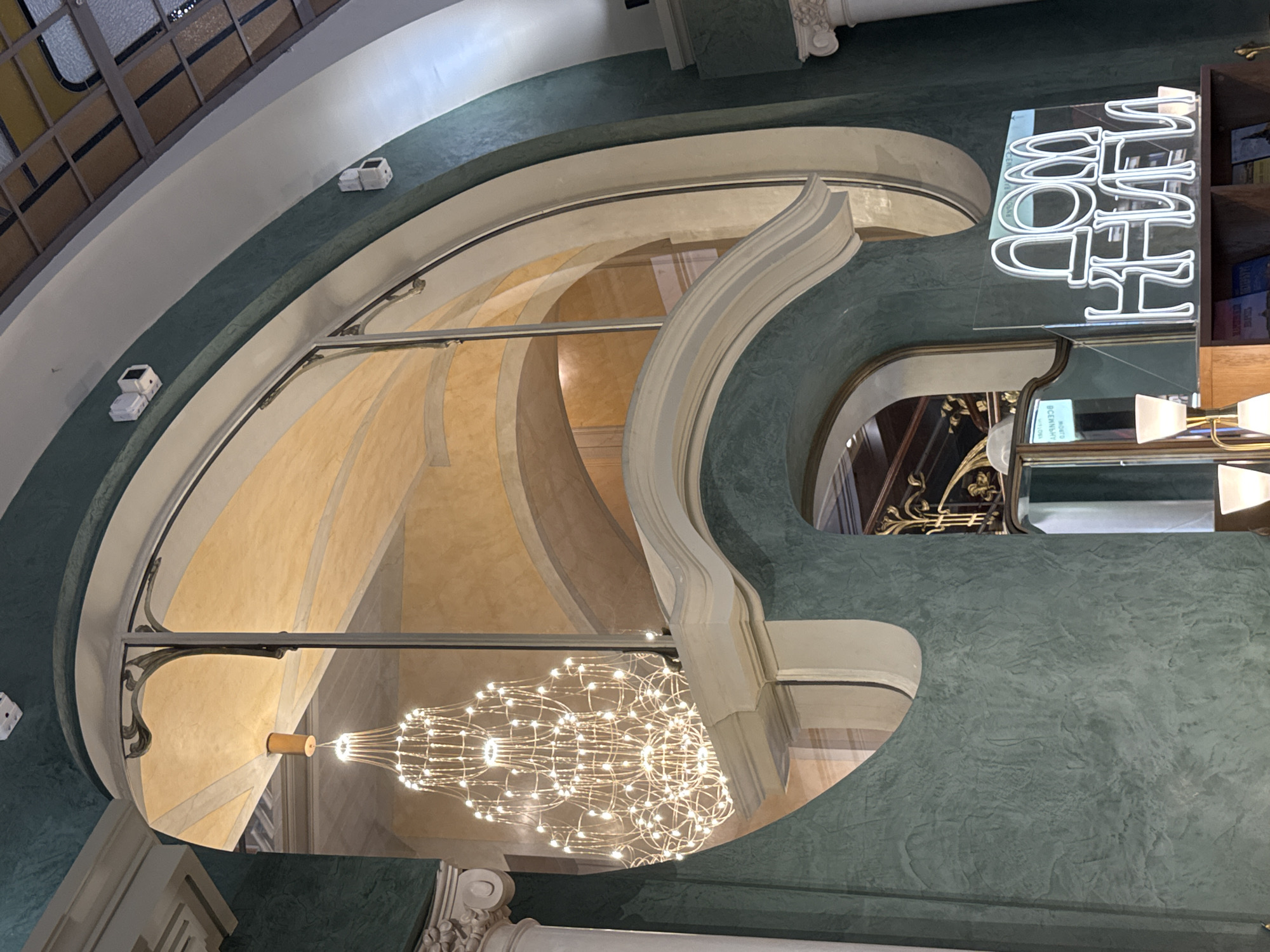 The image shows a view inside the Dom Knigi (House of Books) store, a landmark located at Nevsky Prospekt 28, Saint Petersburg, Russia. The primary focus is on a large, ornate chandelier hanging within a grand, arched architectural space. The walls are a textured, deep teal color, contrasted with cream-colored architectural elements and a warm, pale yellow vaulted ceiling. A large, curved opening reveals further archways and a glimpse of a staircase with a decorative golden railing. On a shelf in the foreground, a neon sign in Cyrillic, spelling "Дом Книги," is illuminated. Books are visible on the shelf below the sign. The overall impression is one of opulent interior design.