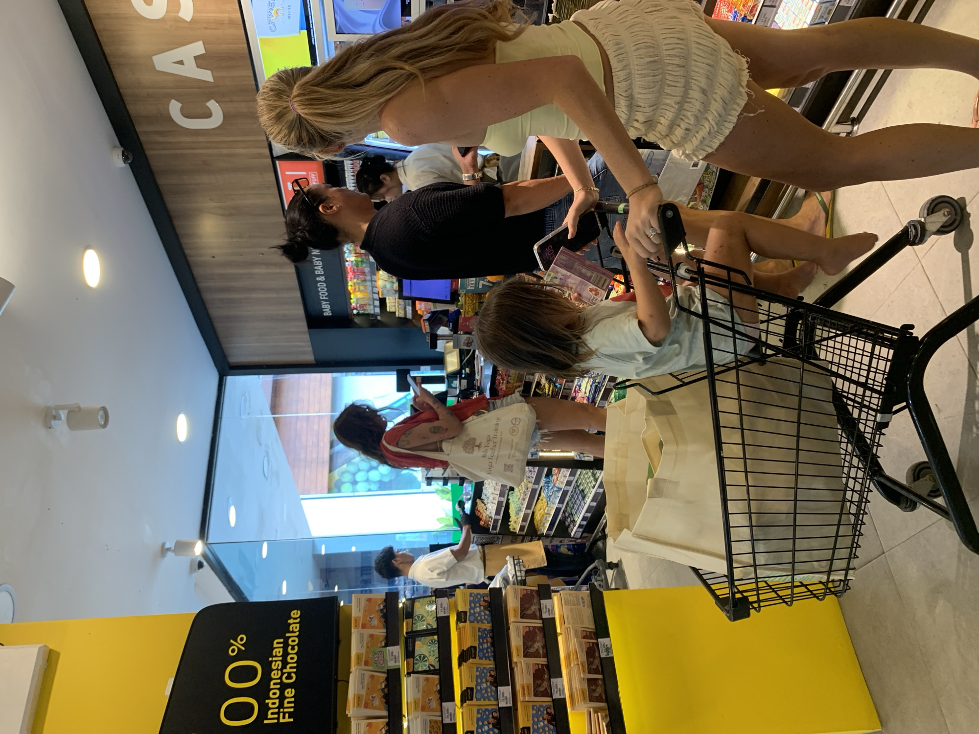 The image shows the interior of a store, possibly a supermarket or grocery store, likely located in Kuta Selatan, Indonesia. A shopping cart dominates the foreground, partly filled with beige shopping bags. A young child is seated inside, holding onto the handle. A woman with long blonde hair and wearing a white ruffled top is either pushing or standing next to the shopping cart. Another woman with dark hair is standing nearby, looking at her phone. In the background, another woman in a red shirt is visible, also looking at her phone. The store has multiple aisles with various products on the shelves, including chocolates and baby food. The natural lighting suggests a daytime setting.