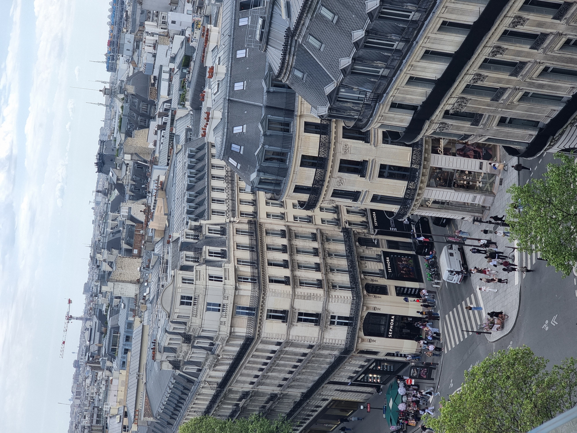 The image depicts an aerial view of a street in Paris, France. The street is lined with tall buildings of Parisian architecture, with ornate facades and multiple floors. The buildings house commercial establishments, including several Sephora stores, as indicated by their signage. Pedestrians are seen walking along the sidewalks and crossing the street at a zebra crossing. A white van is parked on the street. Green trees are visible along the street. In the distance, a construction crane can be seen amidst the cityscape.