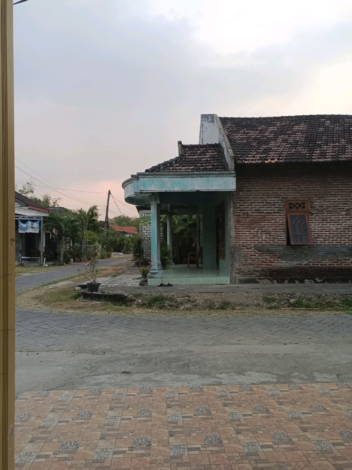 This is a full-frame, eye-level outdoor photograph taken at dusk in the city of Rancang Kencono, Indonesia. The foreground features a patterned tiled patio. Beyond the patio, a paved street runs horizontally, with a narrow strip of dry grass and dirt separating it from the property.
The main subject of the image is a modest, single-story house constructed primarily of red brick with a dark-tiled roof. The house has a covered porch area with a light blue-painted ceiling and white brick pillars. A single window with a wooden frame and a diamond-shaped grill is visible on the brick facade. The house appears to be in a state of disrepair, with some weathering and discoloration on the brickwork.
To the left of the main house, a street continues into the distance, lined with other houses and lush vegetation, including palm trees. A utility pole with overhead wires is prominent in the middle ground. One of the houses on the left has a blue and white sign hanging from its exterior. The sky above is overcast with a soft, diffused light, indicating the time of day is near sunset or sunrise. The overall scene depicts a quiet residential street in a tropical climate. No people are visible in the image.