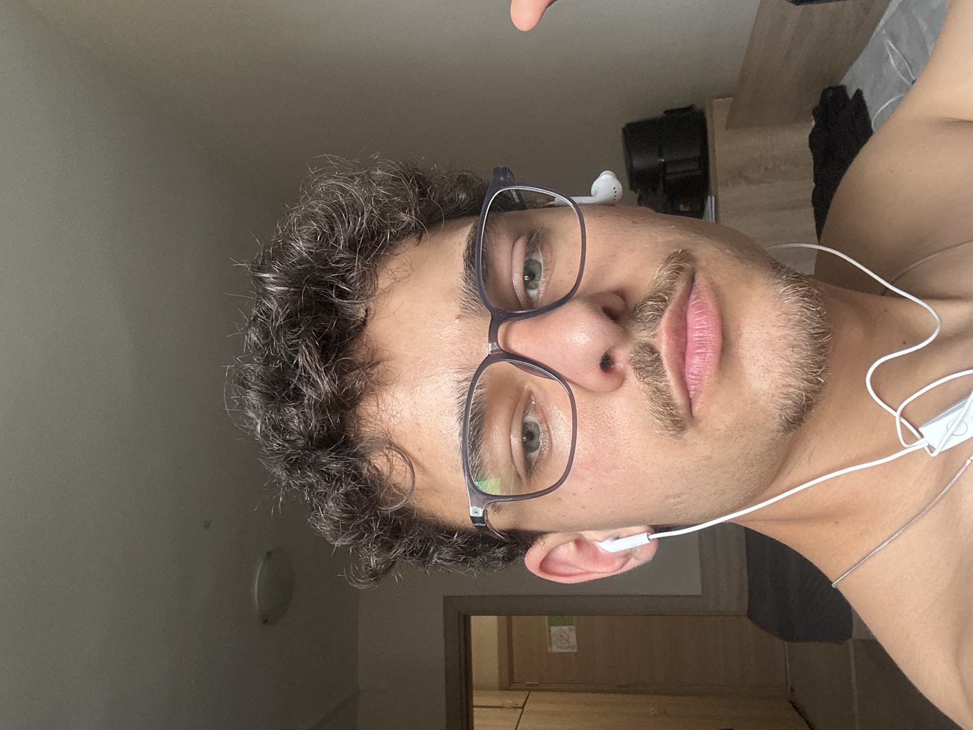 A man with dark curly hair and glasses is facing the camera. He has earbuds in and a silver chain around his neck. The background includes a door frame, a wall with a light fixture, and a shelf with some items on it. There is no information in the image that would indicate this photo was taken in Sofia, Bulgaria.