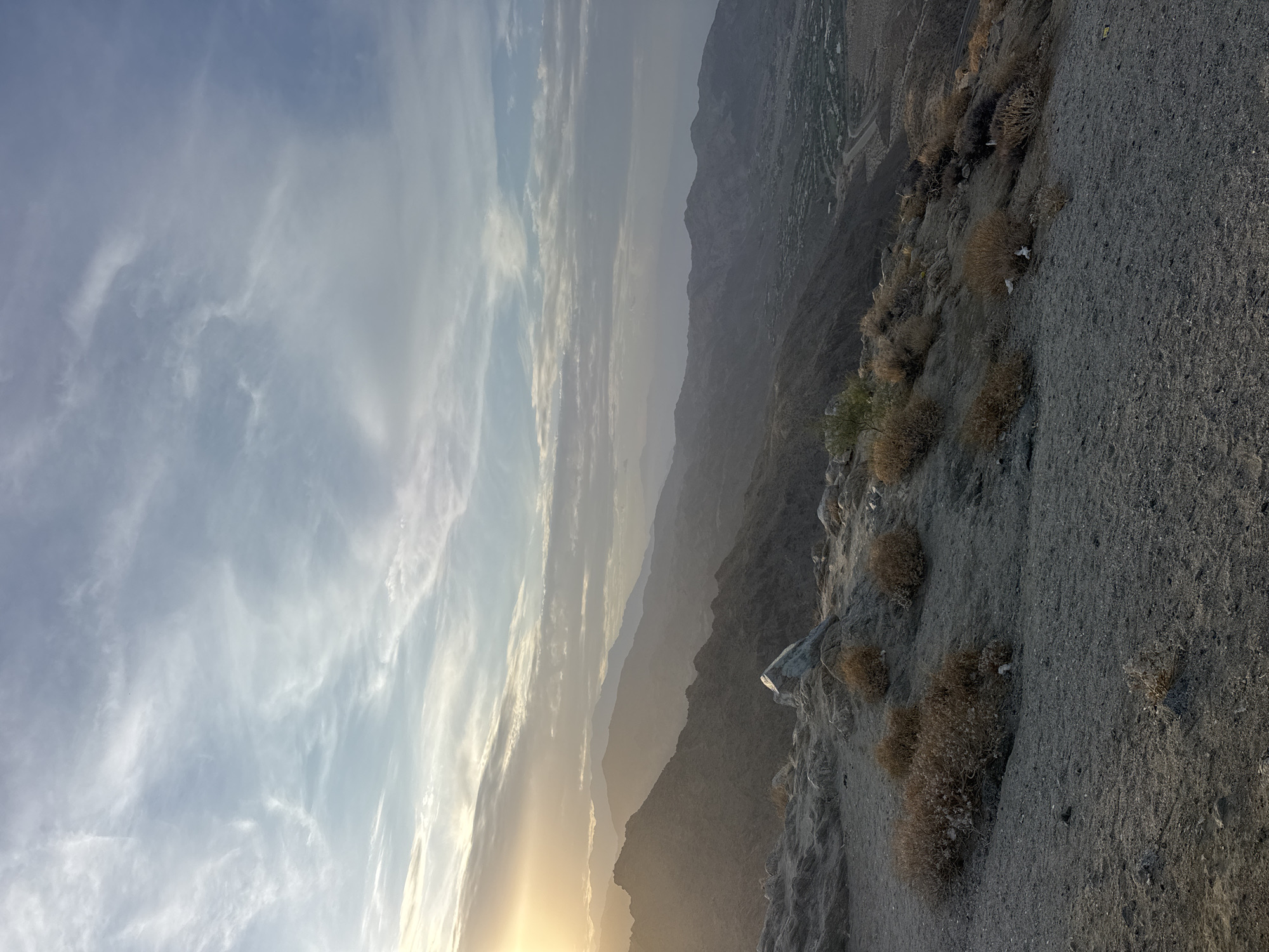 The image captures a wide, elevated view of a mountainous landscape at dusk, with the sun setting in the distance on the left. The sky is filled with wispy clouds, painted in hues of blue, gray, and white, with streaks of golden sunlight breaking through. In the foreground, the rocky terrain is sparsely covered with dry, brown brush and a few larger rocks, including a prominent, angular white rock.
The middle ground is dominated by layers of hazy mountains that recede into the distance, their forms softened by the atmospheric conditions. The setting sun casts a warm, golden glow over the leftmost mountain range, highlighting its rugged texture.
The background reveals a valley with a settlement, featuring buildings and roads, nestled amongst the mountains. A winding road is visible, leading into the valley. The overall impression is one of a vast, serene, and somewhat desolate natural environment. There are no people or animals visible in the image.