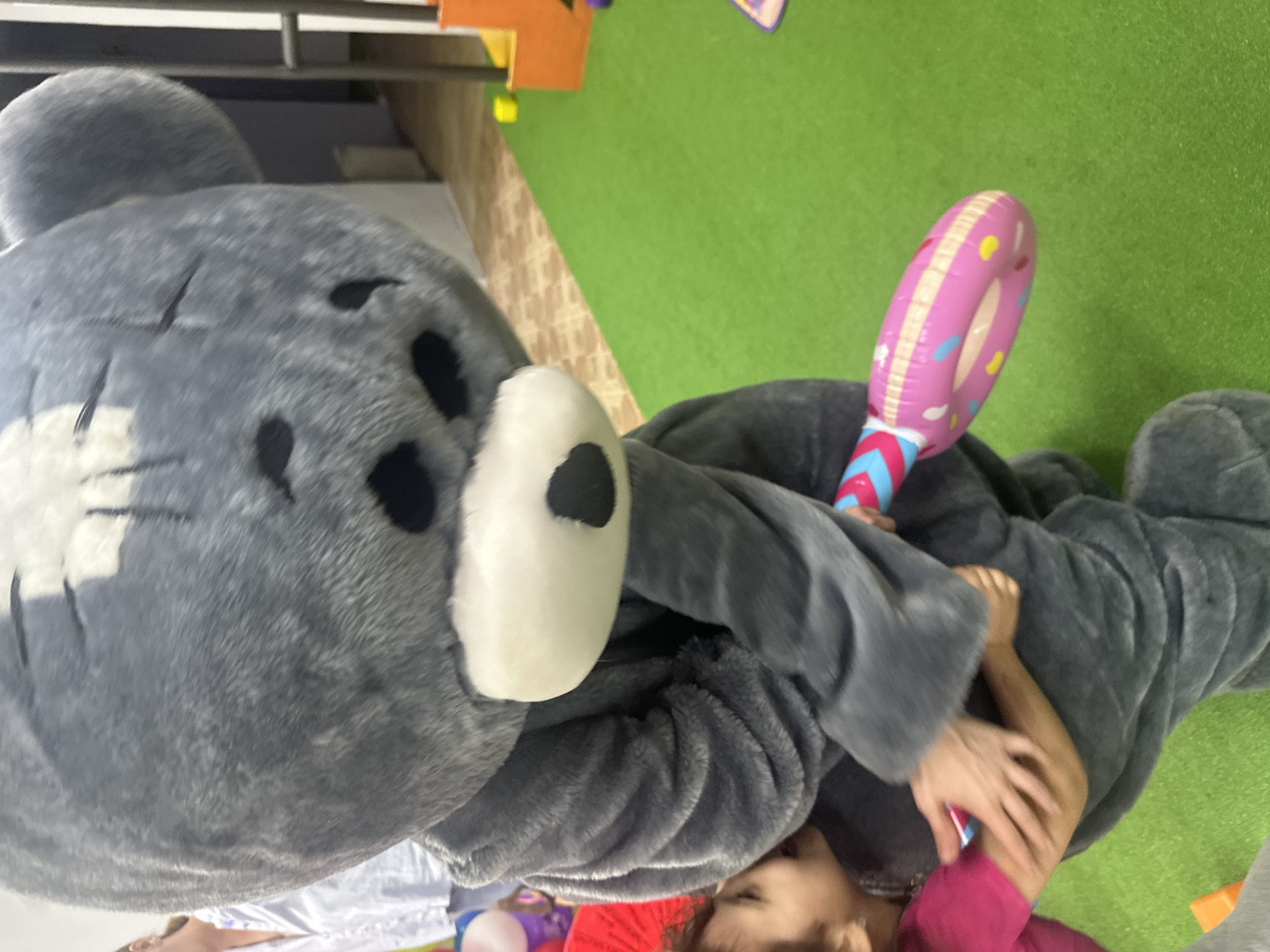 The image captures a playful interaction between a person in a large gray animal costume and another individual, likely a child. The scene is oriented vertically, appearing as if taken from an overhead or unusual angle, with a green mat or carpet making up a significant portion of the background.
The dominant subject is the person wearing the gray, furry costume, which resembles a bear or similar creature. The costume features a large, round head with a white snout, a black nose, and black patches around the eyes. Its fur appears soft and plush. The costume wearer's arms are visible, wrapped around the other person.
The second subject, a child, is partially visible beneath the costume. They are wearing a pink or magenta long-sleeved shirt, and their head, arm, and hand are seen in close proximity to the costume. They appear to be holding a pink inflatable toy shaped like a donut on a stick, adorned with colorful sprinkles in yellow, blue, and white. The stick itself is striped with blue, pink, and white.
The setting appears to be an indoor play area or room. The floor transitions from the bright green mat to a brown patterned surface in the mid-ground. Hints of other play equipment or decor, including orange and yellow shapes and a metal railing, are visible in the background. The overall impression is one of fun and engagement. No text is visible in the image.
