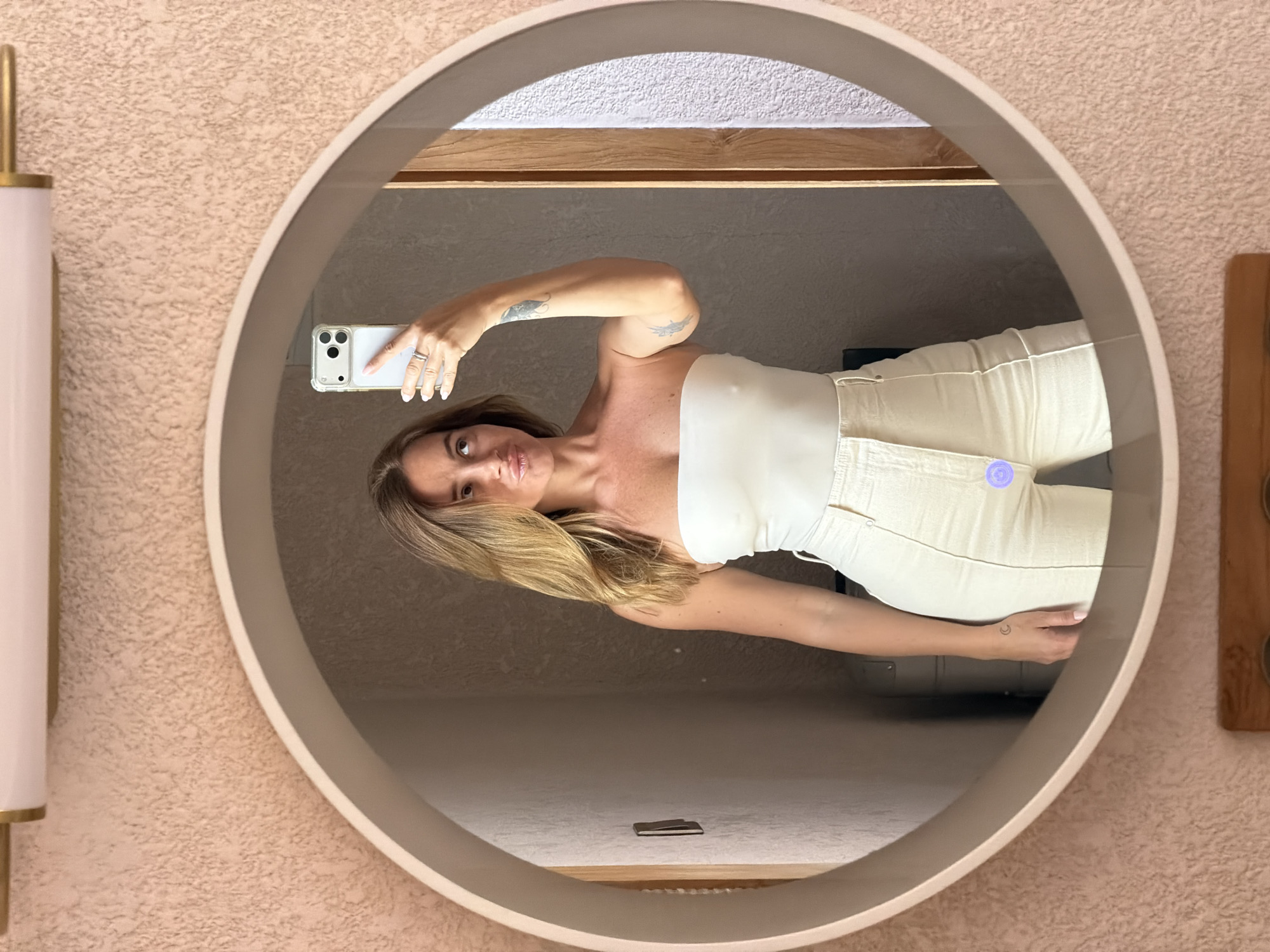 In a bathroom with a textured pink wall, a woman is taking a mirror selfie. She is wearing a white strapless top and cream-colored pants. She is holding a white smartphone in her right hand, capturing her reflection in a large, round mirror. Her hair is blonde and styled loosely around her shoulders. She is making a puckered facial expression. A faint tattoo of a crescent moon is visible on her left forearm. The mirror is framed by a thin, beige border. Above the mirror, a modern light fixture with a pink shade and gold accents is mounted on the wall.