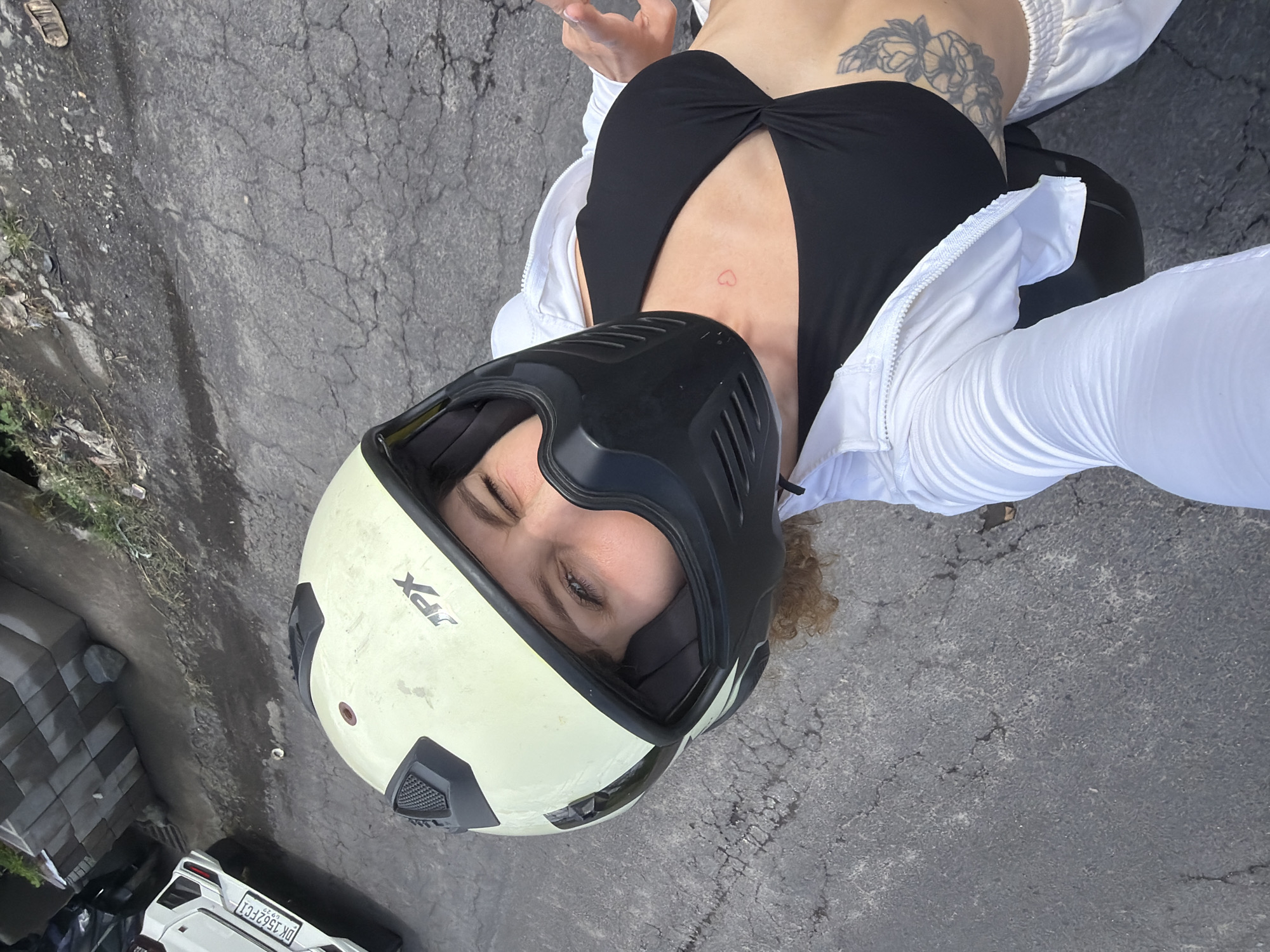 A person wearing a white jacket and a black top with a cut-out is pictured from a high angle, outdoors, in what appears to be a parking lot. They are wearing a cream-colored motorcycle helmet, obscuring most of their face, though their eyes are visible. A tattoo of a flower is visible on their chest.
The scene is set on a cracked asphalt surface. To the left of the frame, there are some stacked gray objects, possibly building materials, and some greenery. A white car with a license plate is also visible in the lower left corner of the image. The license plate reads "DK 1562 FCI".
The time of day appears to be daytime, with bright lighting suggesting sunny weather. The overall mood is casual and perhaps adventurous, given the motorcycle helmet. The person's gaze is directed towards the camera. The image appears to have been taken with a wide-angle lens, as the perspective is somewhat distorted. The location context is Mengwi, Indonesia.