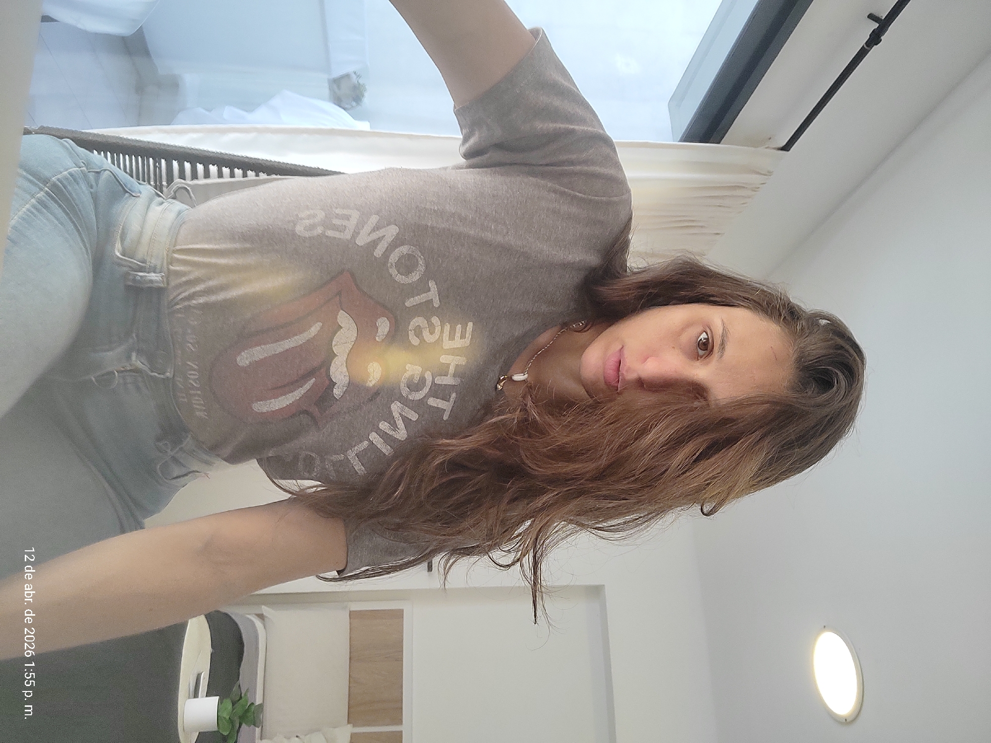A woman with long, brown hair is taking a selfie, facing the camera with her head tilted to the right. She is wearing a grey T-shirt with a Rolling Stones logo and light-blue jeans. She is wearing a necklace.
The background appears to be an interior space with white walls. There is a window with white curtains and a dark frame. A round, ceiling-mounted light fixture is visible. A blurred portion of a balcony or patio can be seen through the window.
The date "12 de abr. de 2026 1:55 p. m." is printed at the bottom left, suggesting this is a digital photograph or screenshot.
There is no specific information in the image to identify the exact location as the city of Buenos Aires, Argentina.