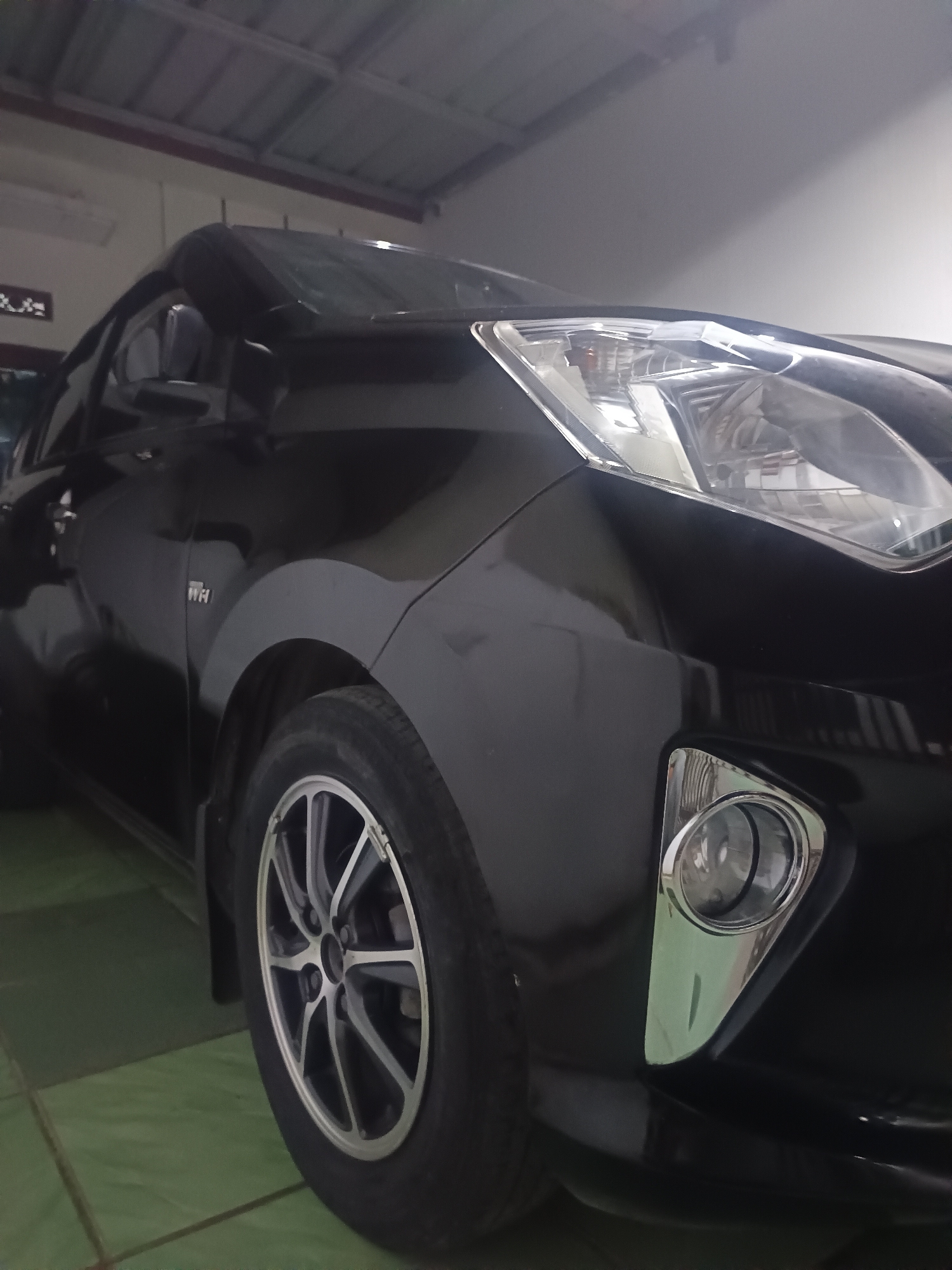 A black passenger vehicle, likely a compact car, is depicted indoors from a low-angle, front-right quarter perspective within the city of Maleber, Indonesia. The vehicle features a reflective black body finish, a clear multi-element headlight, and a chrome-bezeled circular fog light situated in the lower front bumper. A multi-spoke alloy wheel, displaying a two-tone silver and dark grey design, is visible on the front right. An emblem, consistent with "VVT-i", is affixed to the front fender above the wheel arch. The immediate environment includes a floor paved with green square tiles, light-colored walls, and a ceiling with a visible corrugated metal structure supported by white beams. No individuals are present in the scene.
