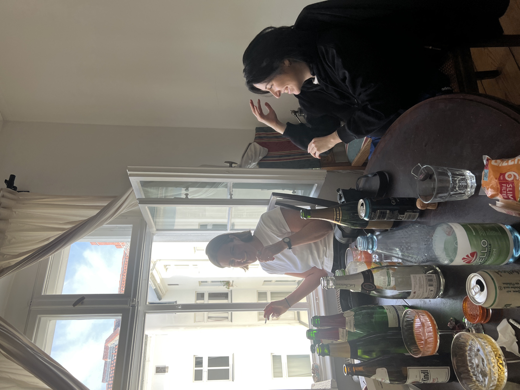 Here is a detailed description of the image:
**Scene Overview:**
The image appears to be taken indoors, possibly in an apartment in Berlin, Germany, as indicated by the presence of "Kindl" beer (a Berlin brewery). The scene is somewhat cluttered, centered around a dark, round table.
**People:**
Two women are present. One is wearing a black sweatshirt and has dark hair pulled back. She is seated near the edge of the table and is gesturing with her hand while smiling. The other woman is wearing a white t-shirt and has shorter, light-colored hair. She is standing near a window and is holding a cigarette.
**Objects:**
* **Table:** A dark, round wooden table dominates the foreground.
* **Beverage Containers:** The table is covered with various empty alcoholic beverage containers including numerous green wine/sparkling wine bottles, several cans (including beer), and a plastic water bottle.
* **Glassware:** Several glasses are scattered around the table.
* **Other items:** The table also holds a pack of slim cigarette filters, a black object possibly a camera or bag, and other miscellaneous objects.
* **Window:** A window with multiple panes is visible in the background, looking out onto a courtyard.
* **Curtain:** A sheer, white curtain is partially drawn over the window.
* **Furniture:** A wooden chair is visible behind the woman in black.
* **Building:** The building across the way is white.
**Actions and Interactions:**
The two women appear to be engaged in a conversation. The woman in black is gesturing and smiling, and the woman in white is smoking and looking in her direction. They appear to be enjoying a relaxed social gathering, possibly after a party or celebration, given the number of empty alcoholic beverage containers on the table.
**Environment:**
The room seems to be well-lit, possibly with natural light coming through the window. The setting suggests a casual, lived-in apartment in a city environment. The external architecture visible through the window appears to be typical of many older buildings in Berlin.