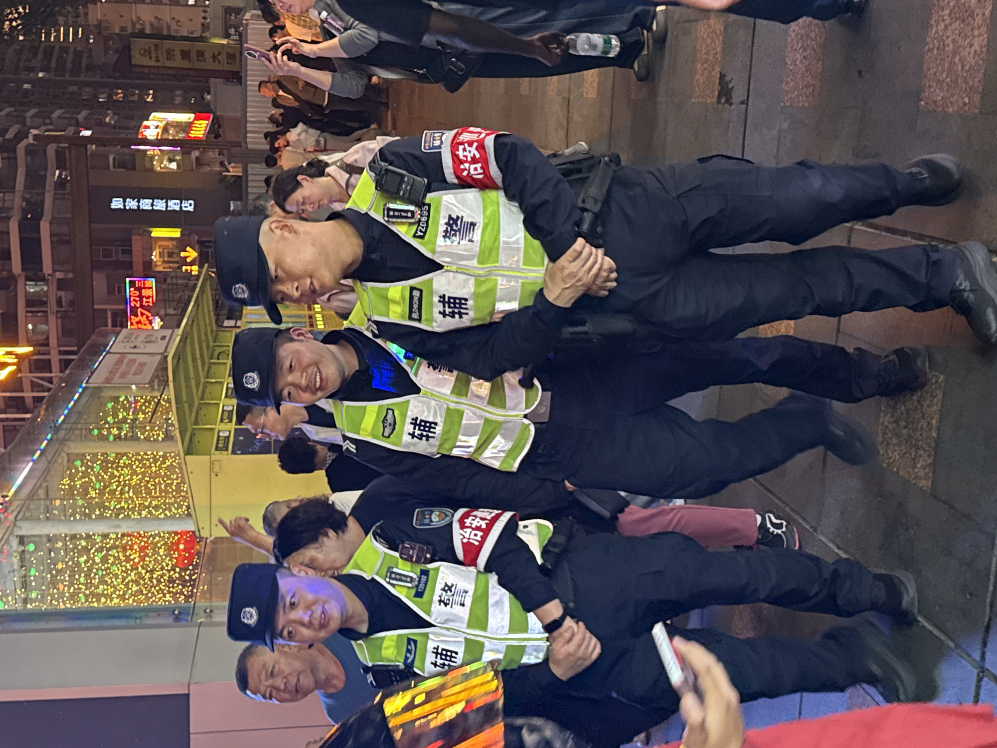 This image depicts four uniformed officers, presumably Chinese Auxiliary Police ("辅警"), standing on what appears to be a city street at night. They are wearing dark blue uniforms, black caps with emblem and neon green reflective vests with Chinese characters "辅警 (Auxiliary Police)" and "执勤 (On Duty)" written on them. One officer has the identification number YZ0695 on their vest and another has YZ0192. One officer has "执ZHIQIN勤" on the vest. Some officers are wearing armbands with the word "治安" written on them.
The setting is an urban area, with numerous buildings, illuminated signs, and other people in the background. The Chinese characters on the signs suggest the location to be in Yuzhong District, China. The sign on the left of the officers read "如家商旅酒店", or Home Inn Hotel. The LED sign above this says "270℃ 江景" and the characters "農大厦". The lighting is mainly provided by streetlights and the signs. Many people are milling around the officers.