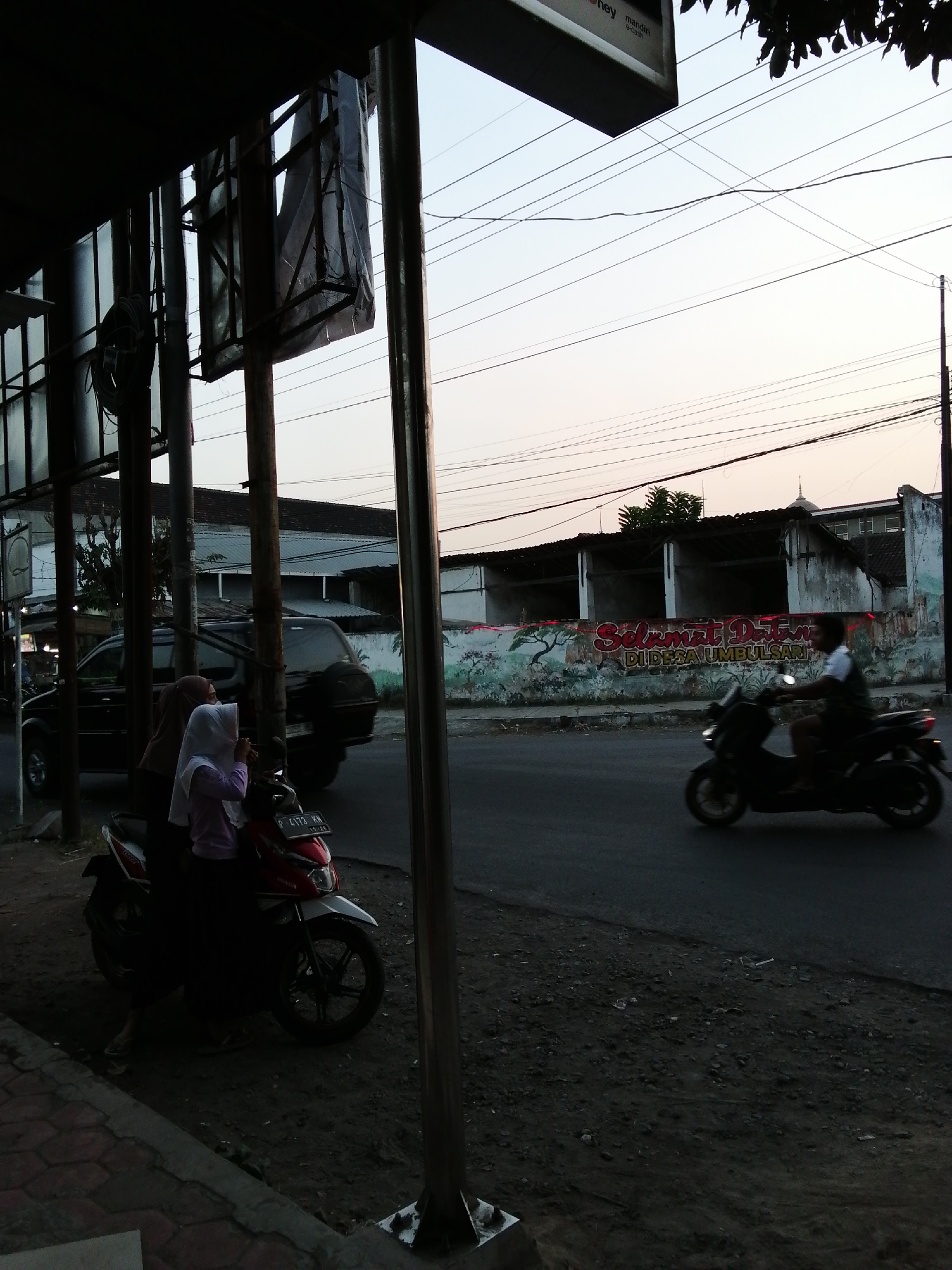 An exterior shot shows a street scene in the late afternoon or early evening in an unknown city. In the foreground, two veiled women stand next to a red and white scooter parked on the side of the road. One woman wears a brown headscarf and a long dress, while the other wears a white headscarf and a pink shirt. A black SUV is parked on the street behind them, partially obscured by a vertical pole. The road is paved, and the ground on the side of the road is covered in dirt and small stones. In the background, a long, low building with a mural on its wall is visible. The mural depicts trees and a procession of animals. A person on a black scooter is riding down the street to the right of the frame. Overhead, a tangle of electrical wires crisscrosses the sky. The sky is a pale gradient of blue and pink, suggesting the sun is setting. The scene is dimly lit, with shadows cast by the overhead structures.