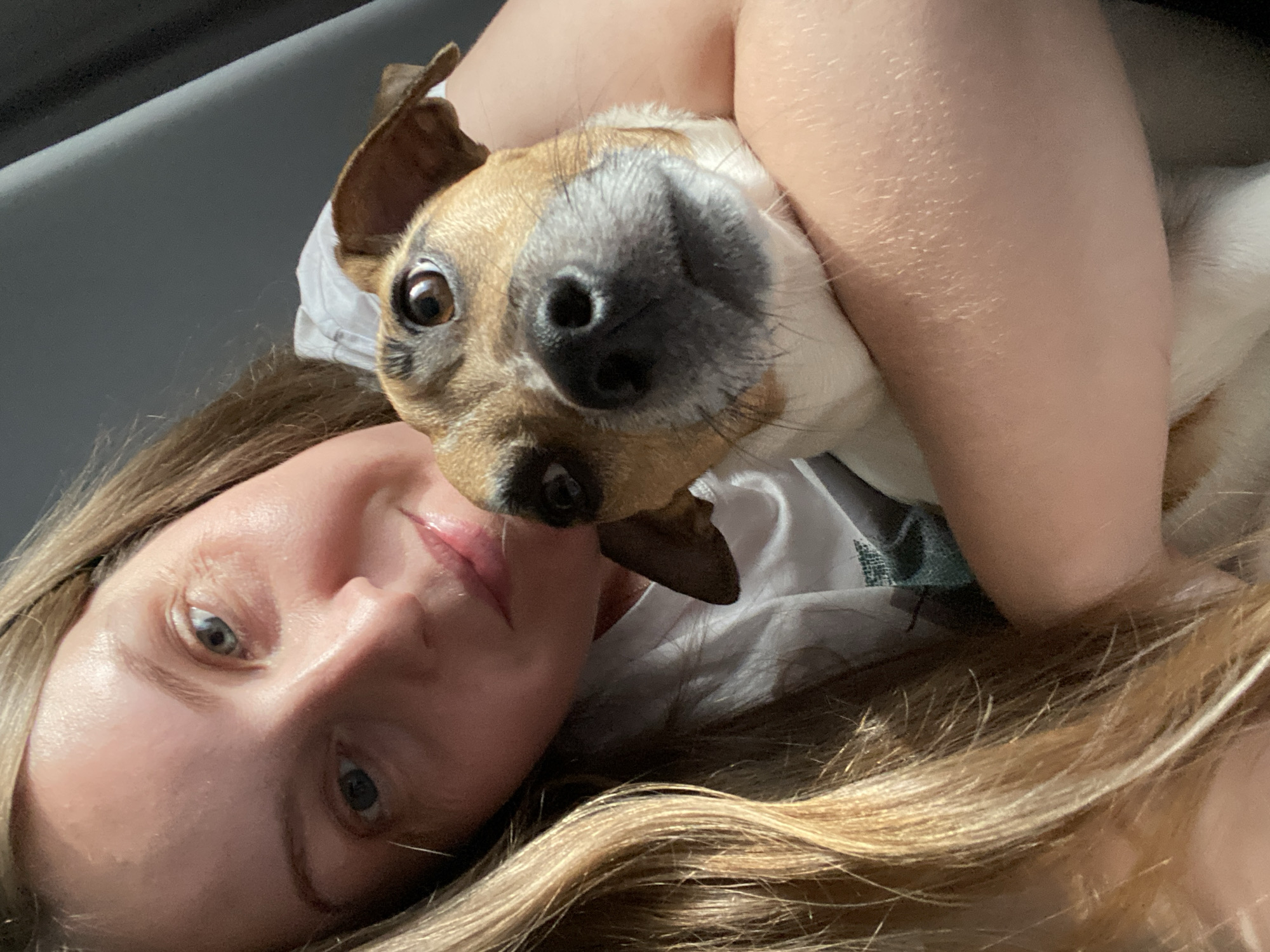 A selfie-style close-up image shows a fair-skinned person with blonde, wavy hair lying on their back. The person has light eyes and is looking directly at the camera. Cradled in the person's arms is a dog, mostly white with brown markings around the head and ears. The dog's large, dark nose is prominently featured. The background appears to be a simple, muted gray surface, possibly a bed or couch.
There is no information to suggest the image was taken in Noviy, Russia.