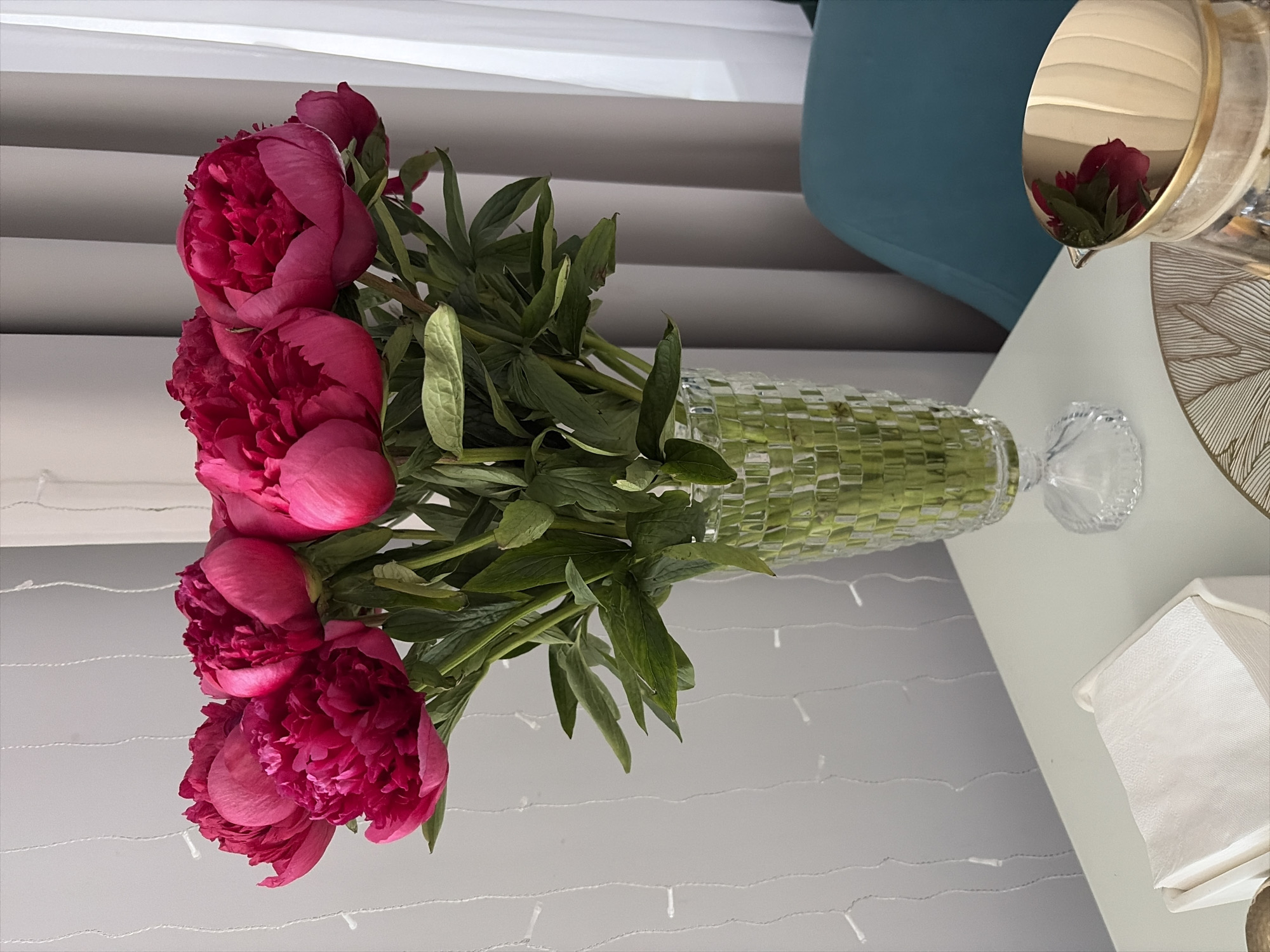 In a room with light grey walls and vertical drapes, a clear glass vase filled with a bouquet of vibrant pink peonies stands on a white table. The vase has a patterned texture. Next to the vase, a white napkin holder with folded white napkins is visible. In the foreground, a decorative plate with a brown line pattern is partially visible. To the right of the plate is a silver pitcher with a gold rim, reflecting a dark red flower and green leaves. A teal-colored chair is visible in the background. String lights hang on the wall. There is no indication that this scene is located in Moscow, Russia.