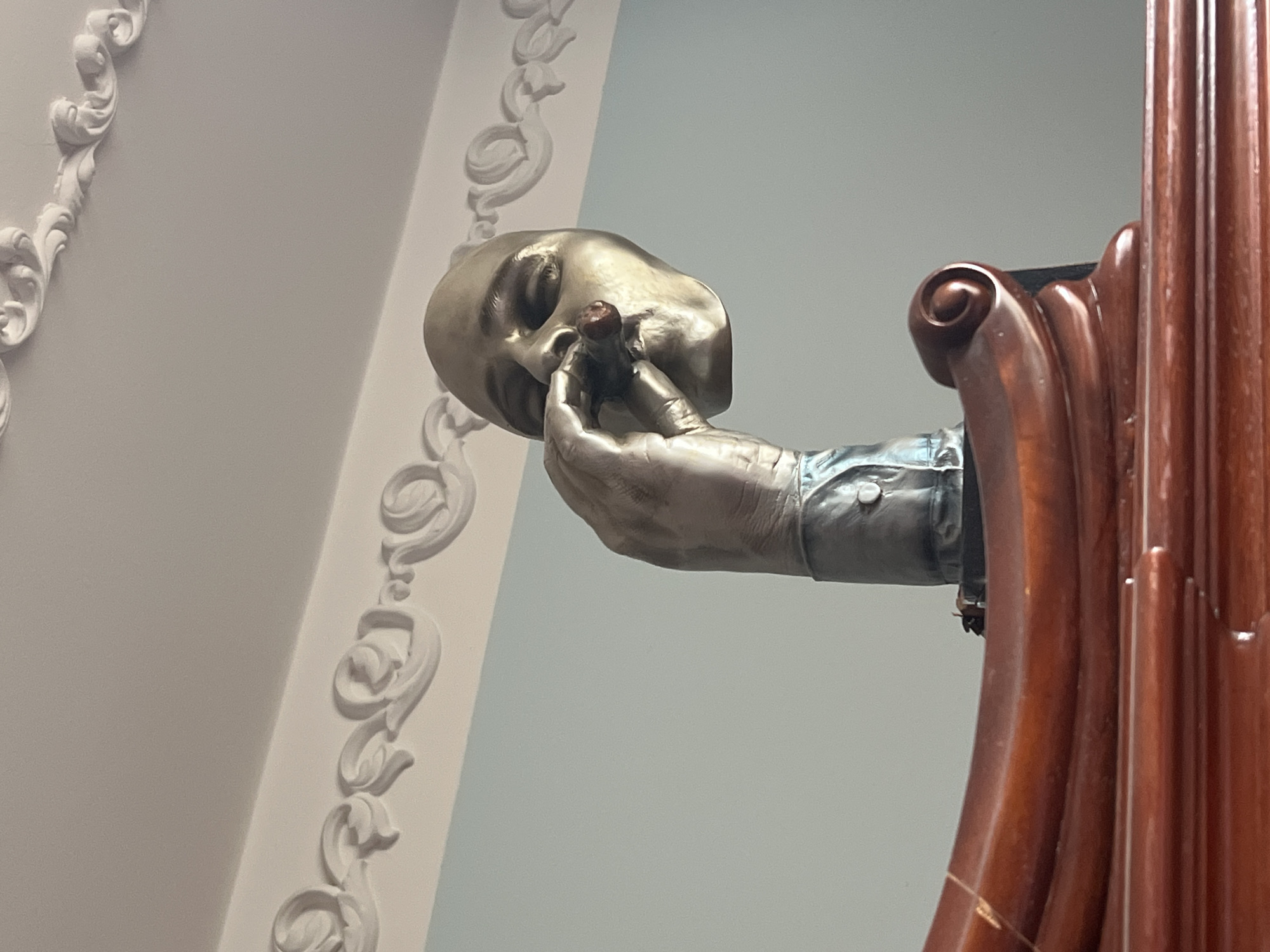 The image shows a close-up of a statue of a head and hand holding a cigar, situated on top of a polished wooden surface. The statue's head is tilted back, with its eyes closed. The hand is positioned near the mouth, holding the cigar with its fingers. The statue appears to be made of a metallic material, possibly bronze or gold-toned. The wooden surface is elaborately carved with a curved design. The background consists of a light blue wall and white ornate molding along the ceiling. The overall setting suggests an indoor environment, possibly a museum, gallery, or a wealthy individual's home. The image does not provide any visual cues to pinpoint its exact location in Sochi, Russia.