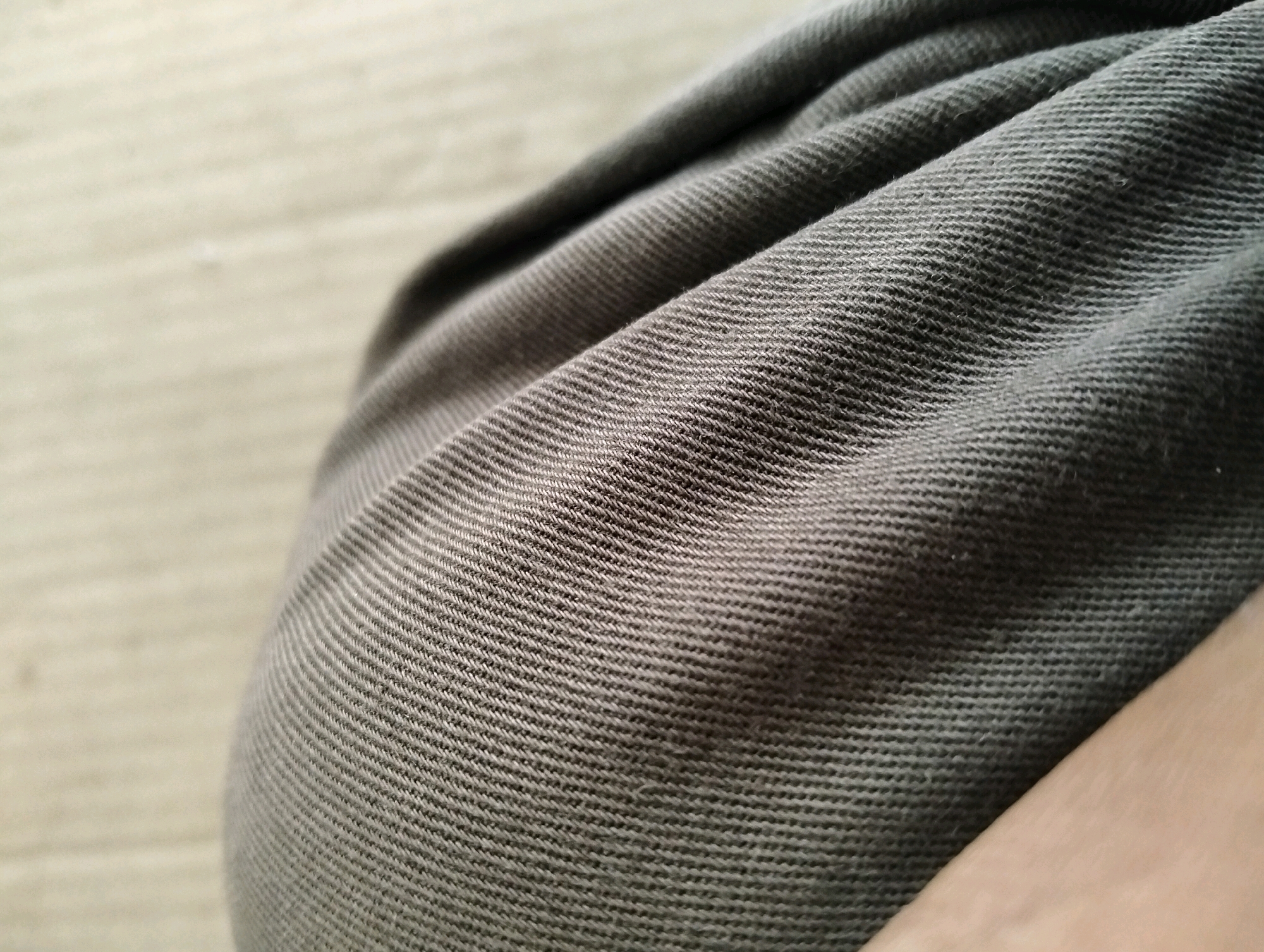 A close-up, shallow depth of field shot focuses on the texture of dark green fabric, possibly denim or a similar twill weave. The fabric is creased and folded, creating a series of parallel lines and shadows that give it a textured, three-dimensional appearance. To the left of the fabric, a lighter, out-of-focus background with a subtle striped pattern is visible, suggesting a neutral surface like a wall or a piece of furniture. The lower right portion of the image shows a sliver of a light-skinned hand or arm, also out of focus, which seems to be the source of the folds in the fabric. The scene is indoors, with even lighting that highlights the texture of the material. There is no discernible activity or interaction depicted, only a static arrangement of fabric and a hand. The location in Tanggulangin, Indonesia, is not visually represented in the image itself; it is contextual information provided separately.