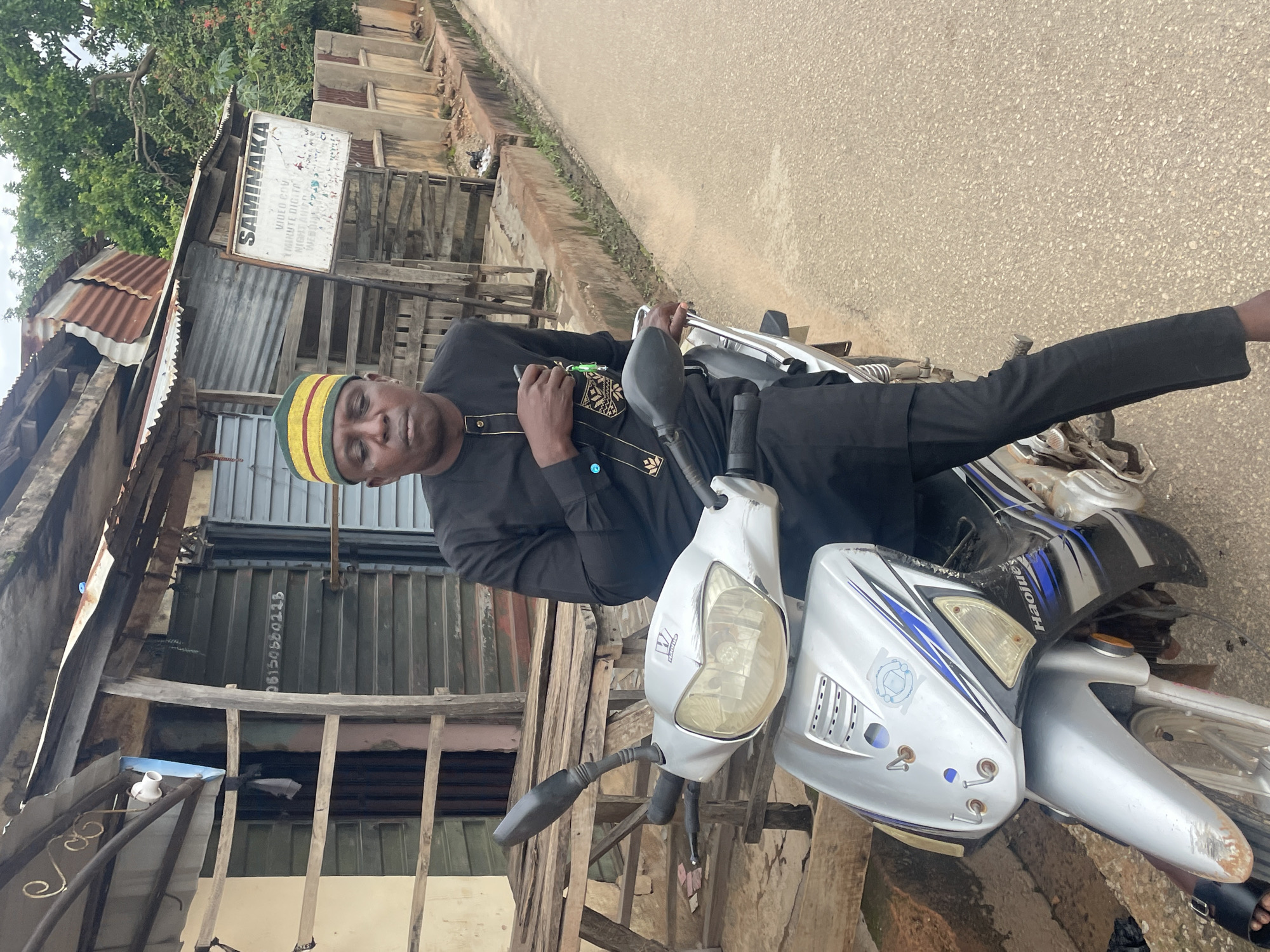 A man wearing a black traditional outfit and a green and yellow striped cap sits on a silver motorcycle. He is holding a dark object, possibly a phone, in his left hand. The motorcycle is parked on an unpaved road. In the background, there is a weathered wooden structure with a corrugated metal roof. A sign attached to the structure reads "SAMINAKA VIDEO CUN Y.MINTEDIGITT NORT NIGHT AMBA YEROWA OK." The scene appears to be in a rural or semi-urban area in Jos, Nigeria. The lighting suggests it is daytime, and the sky is overcast, indicating possible cloudy weather. The overall atmosphere seems casual, with the man posing for the photo. The motorcycle has blue and silver decals and appears to be used. The buildings in the background are simple and constructed with readily available materials like wood and metal, typical of many communities in Nigeria. The road surface is a mix of gravel and dirt.