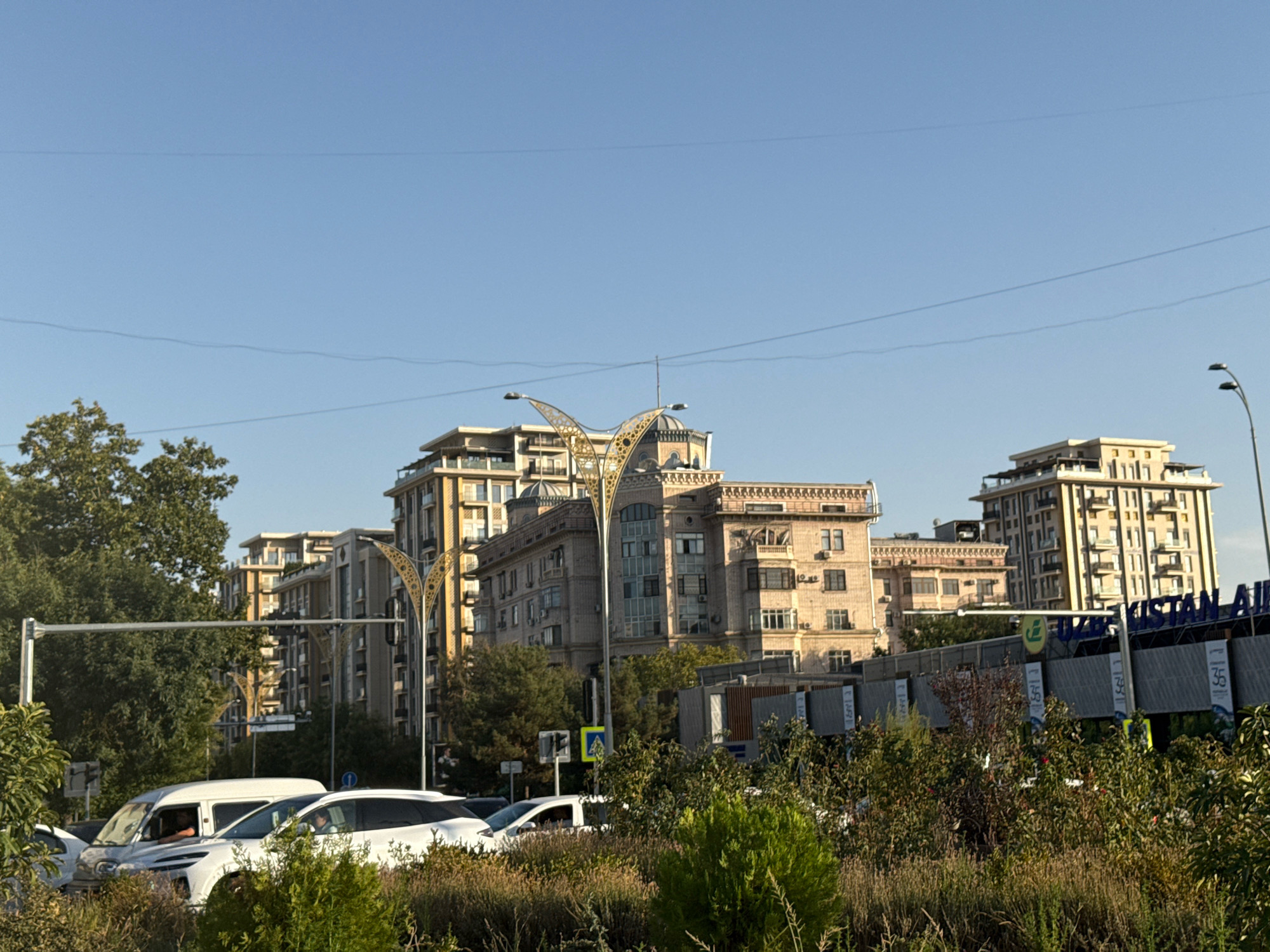 The image depicts a street scene in Tashkent, Uzbekistan, likely in the vicinity of Tashkent International Airport. In the foreground, there is lush greenery and bushes, with the tops of several cars visible, including a white van and a white SUV. The vehicles are stopped, suggesting traffic or a stop sign. In the midground, a traffic light is visible, along with a street sign. Behind the traffic light, there are several modern, multi-story buildings with distinct architectural styles, characterized by beige facades, numerous windows, and balconies. One building features a prominent, decorative golden archway. Further in the background, more buildings and trees are visible under a clear blue sky. A large sign for "UZBEKISTAN AIRWAYS" is partially visible on a structure in the background. The overall impression is a bustling urban environment with a mix of modern architecture and natural elements.