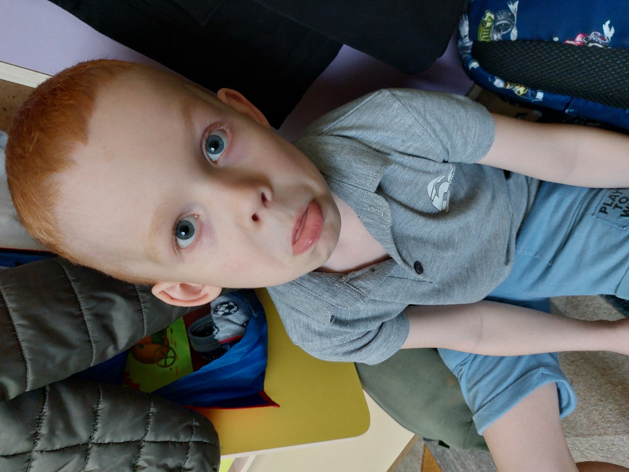 A boy with red hair and blue eyes is looking upwards at the camera with his tongue slightly sticking out. He is wearing a gray short-sleeved shirt and light blue shorts. He is surrounded by various objects, including a dark-colored jacket, a blue backpack, and a yellow and green surface. The location is indoors. There is no information available to verify this image was taken in the city of Chaikovsky, Russia.
