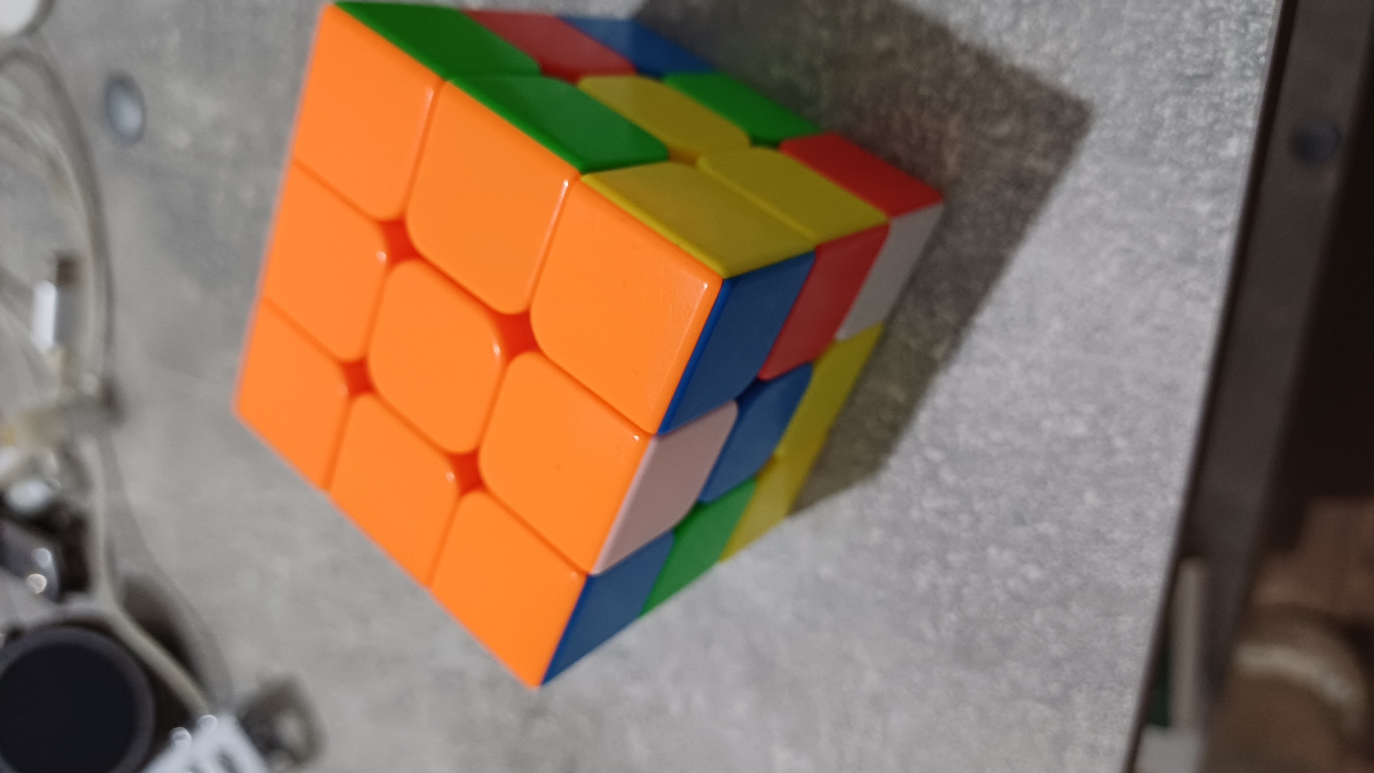 A 3x3 Rubik's Cube is positioned on a grey, textured surface. The cube's visible faces show several colors: orange on the top and right sides, green on the right side, blue on the front and right sides, yellow on the bottom right, and a light pink cube on the front. The surface appears to be a tabletop, with some out-of-focus objects in the background, including what looks like electronic devices and cables. The lighting suggests an indoor environment. The image does not provide any information about the city of Nagornoye, Russia.