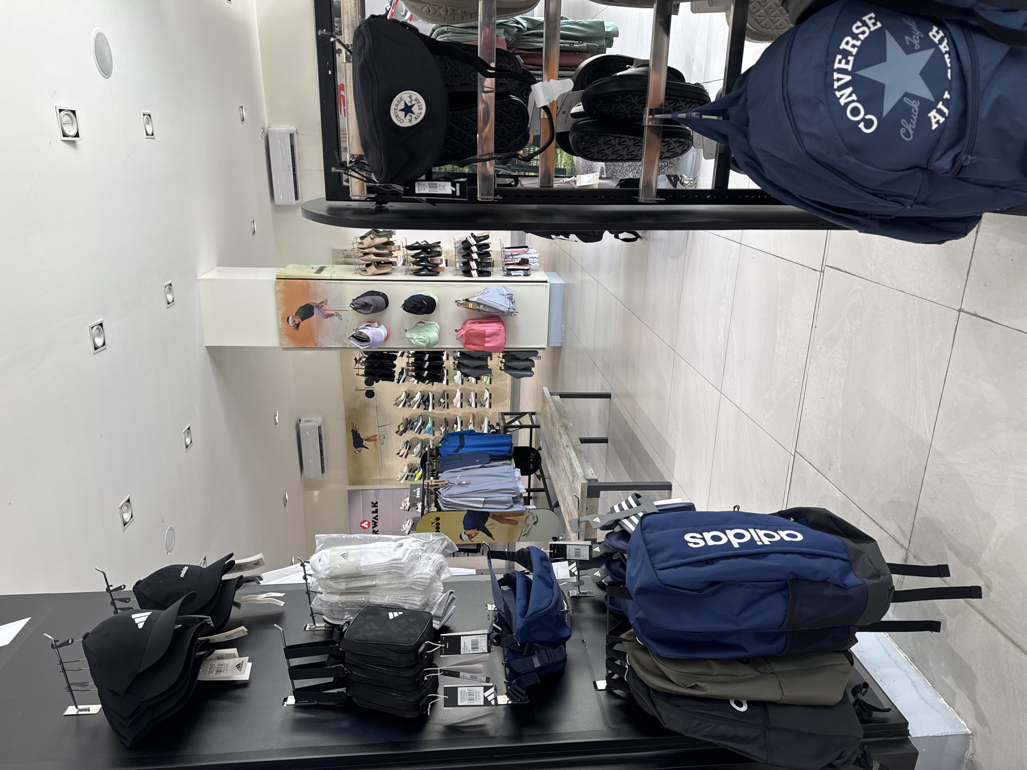 The photo shows an elevated view of a retail store in North Kuta, Indonesia. The store sells clothing, shoes, and accessories. There are displays of Adidas hats, Converse backpacks, and Adidas backpacks. The store has bright, white walls and tile floors. There is no one visible in the photo.